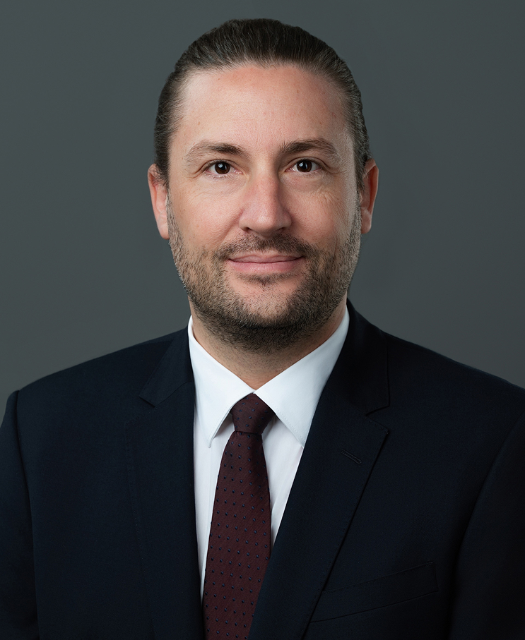 A professional headshot of a man with dark hair, light skin, wearing a dark suit, white shirt, and maroon tie, smiling slightly against a gray background.