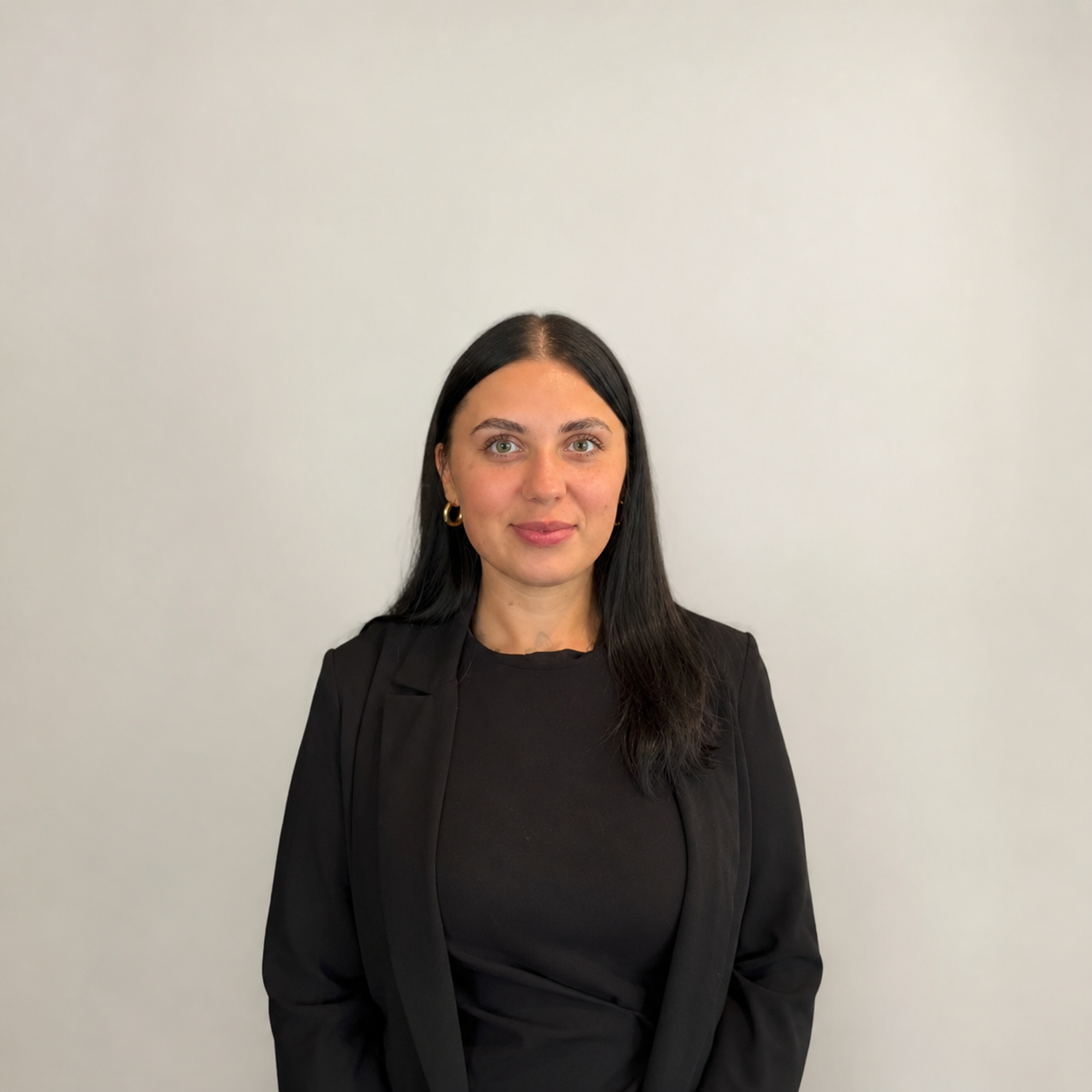 A woman with long black hair, light skin, and green eyes, wearing gold hoop earrings, a black top, and a black blazer, standing against a plain white wall.