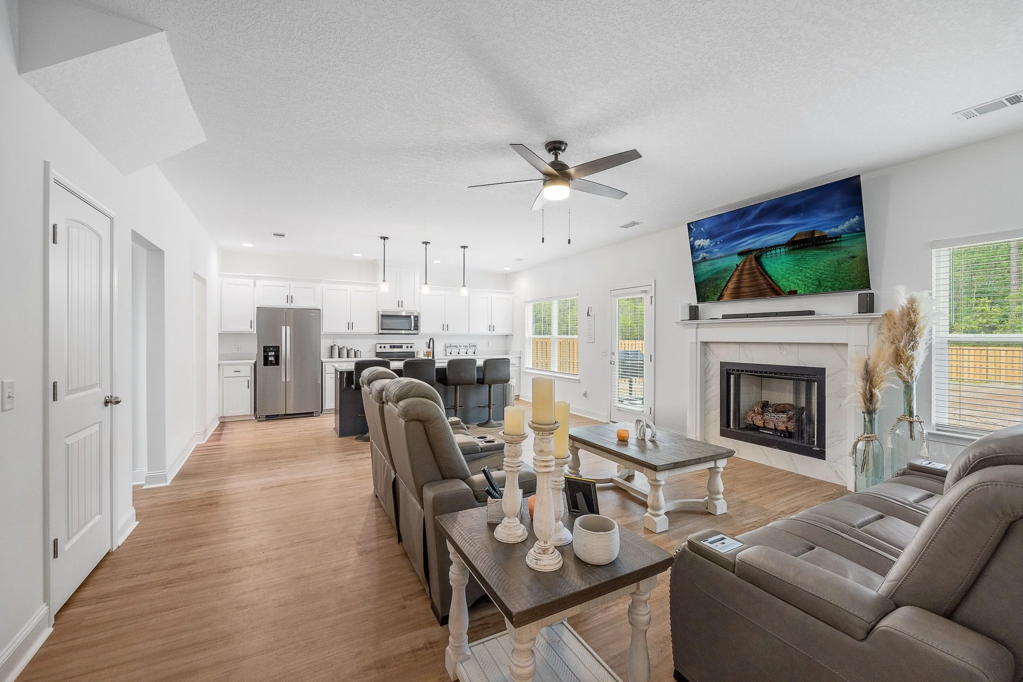 Open-concept living room and kitchen with white cabinets, stainless steel refrigerator, ceiling fan, large television above fireplace, beige recliner chairs, gray sofa, wooden flooring, and large windows letting in natural light.