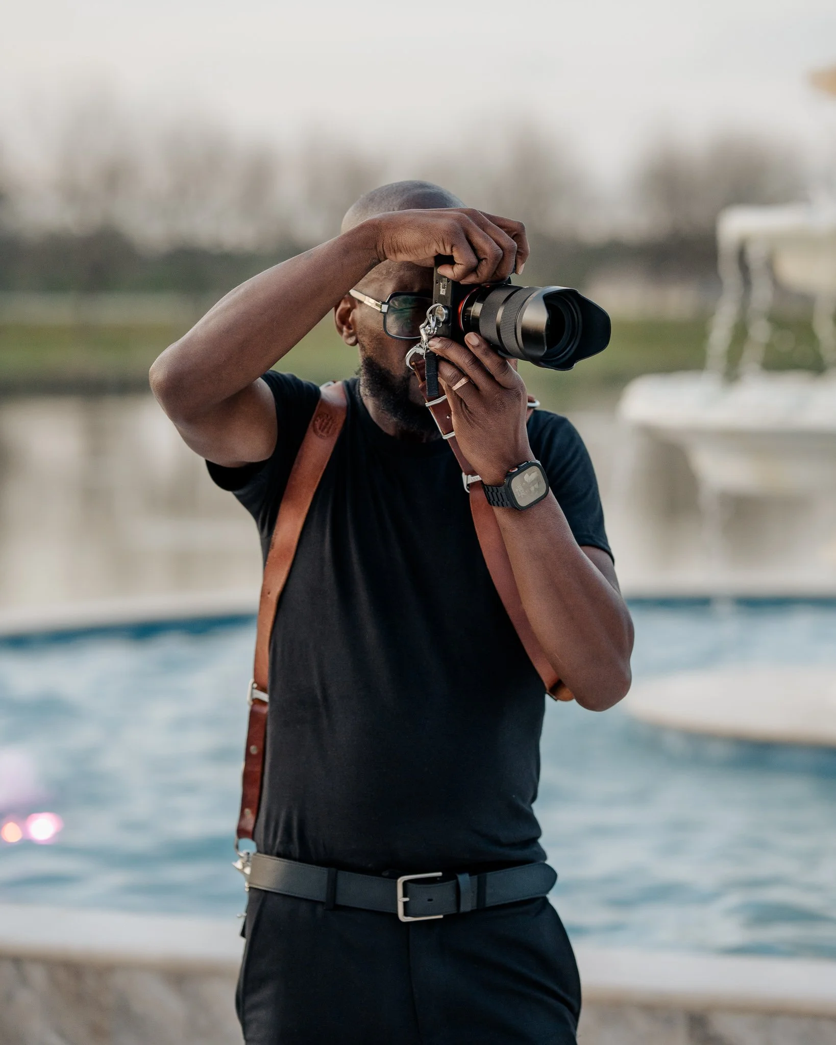 A man taking a photo with a camera outdoors near a body of water, wearing a black shirt, glasses, and a watch, with a brown strap backpack.