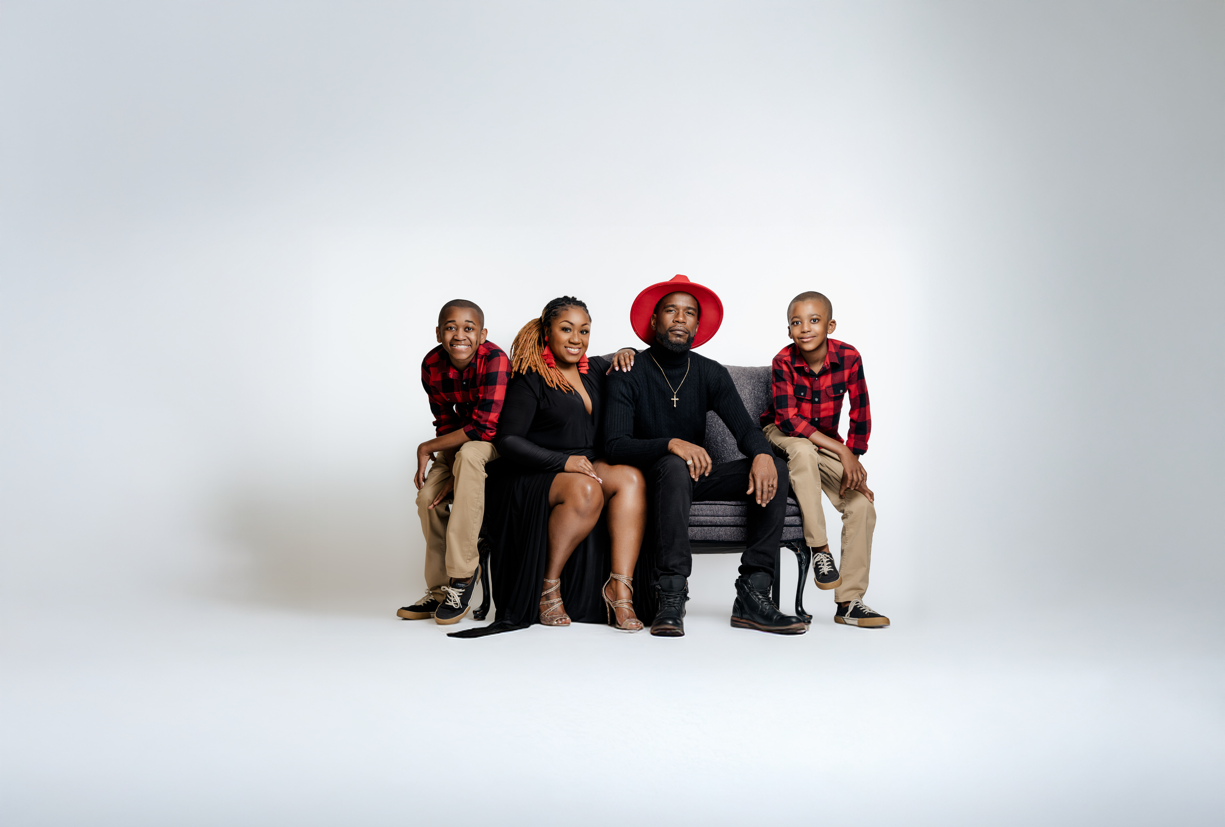 A family of five posing on a bench against a plain white background. The family consists of a man, woman, and three children. The man is wearing a black sweater, black pants, black boots, and a red wide-brimmed hat. The woman is dressed in a long black dress and silver heels. The children are dressed in matching red and black plaid shirts and khaki pants. The family appears happy and relaxed.