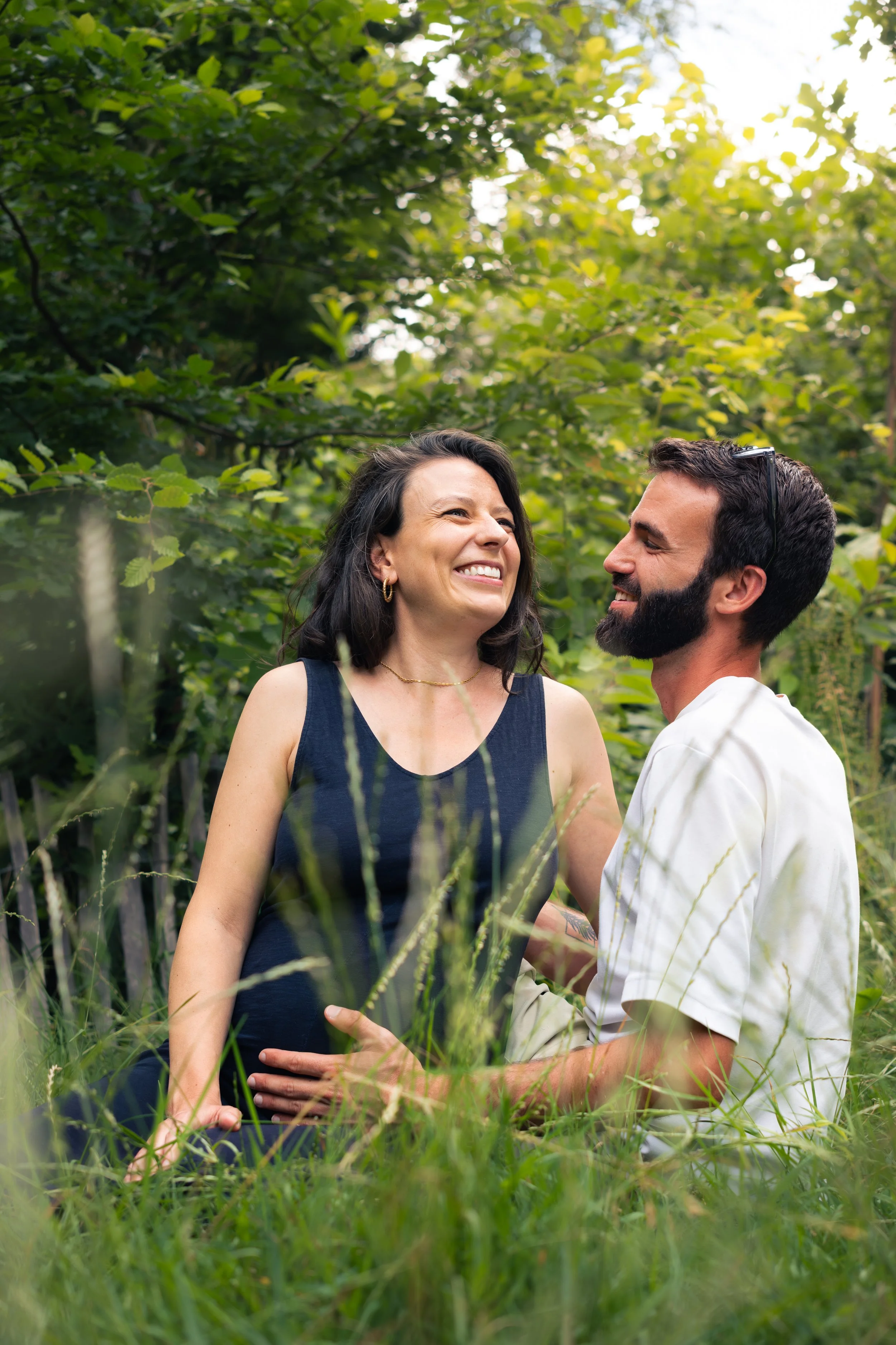 Un homme et une femme enceinte souriants, debout dans un environnement boisé, partageant un moment de complicité dans la nature.