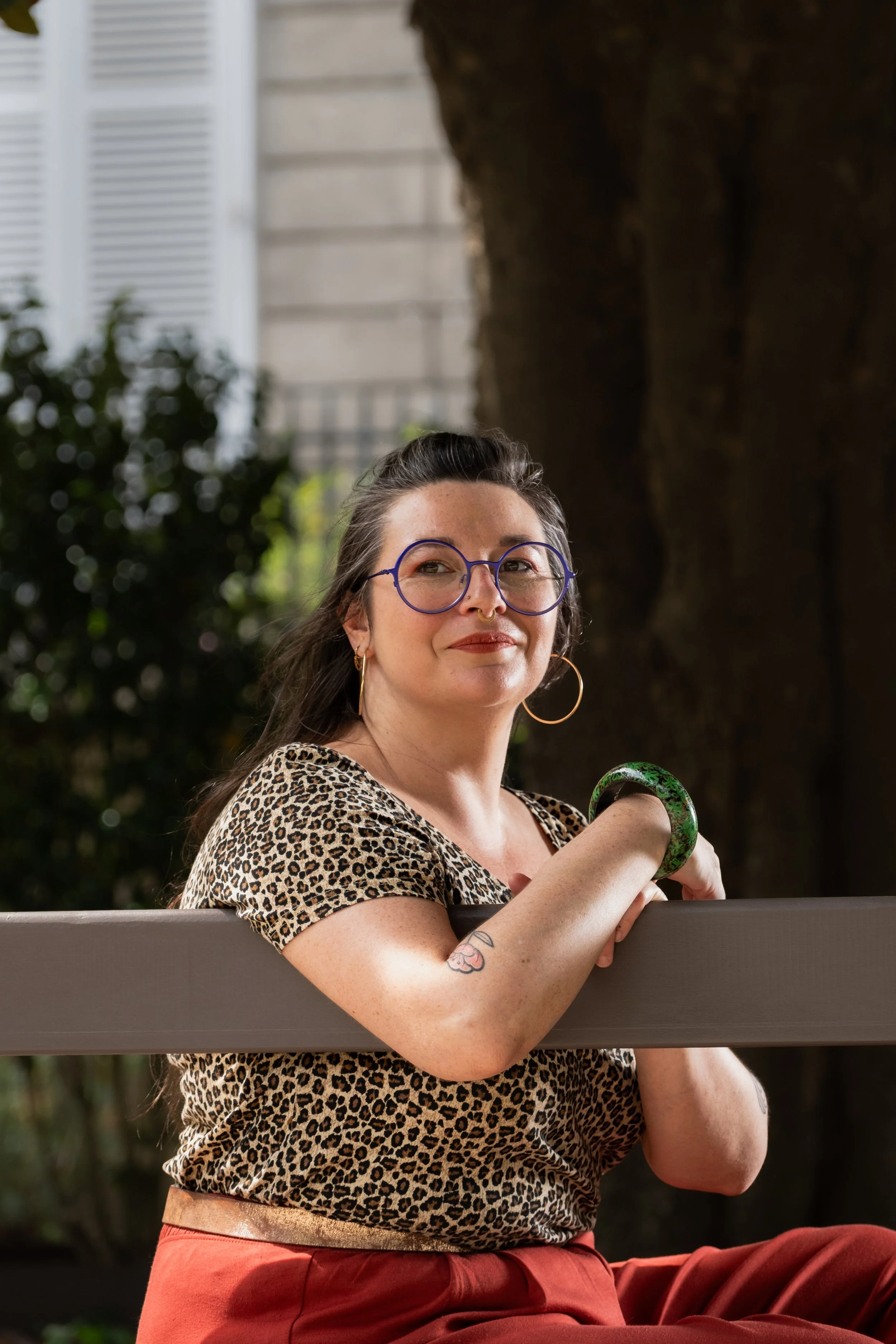Une femme portant des lunettes rondes, un haut à motif léopard, et de grands boucles d'oreilles, assise dehors en tenant une barre ou une rampe, avec un fond flou d'arbres et d'un bâtiment