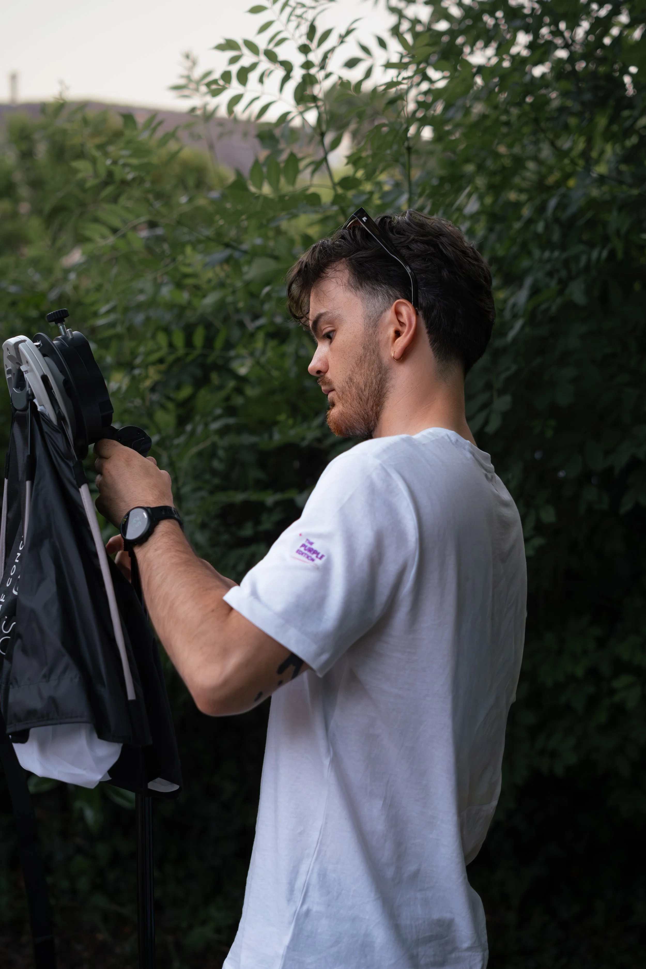 Un homme jeune, avec des cheveux bruns, portant une t-shirt blanc, ajuste un équipement pro de photographie ou de vidéo en extérieur : une softbox avec un flash pour une lumière de qualité.