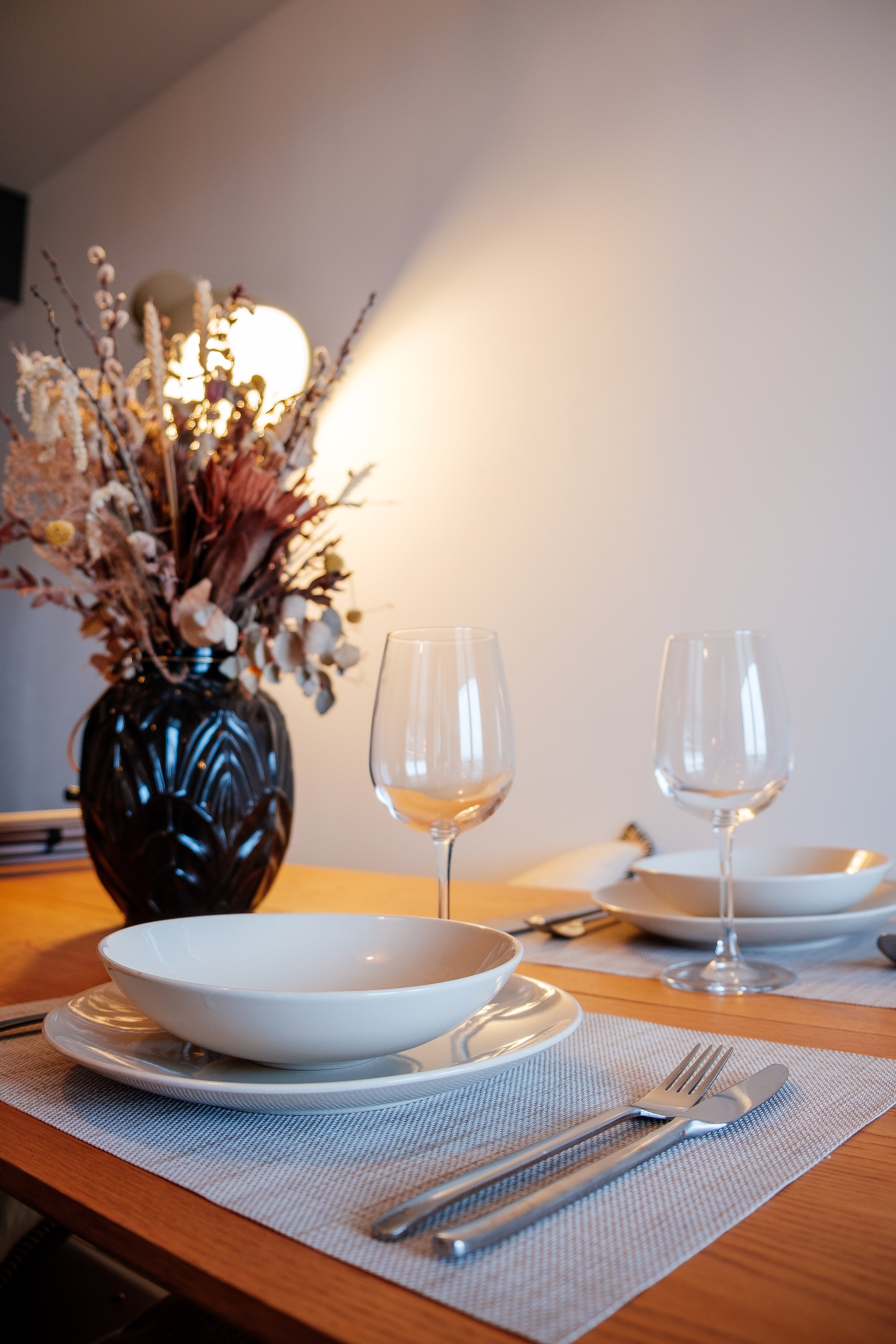 Une table élégante avec des assiettes blanches, des verres à vin vides, une coupe à vin, une fourchette et un couteau en acier inoxydable. En arrière-plan, il y a un vase noir contenant des fleurs séchées aux tons neutres, et une lumière diffuser cré