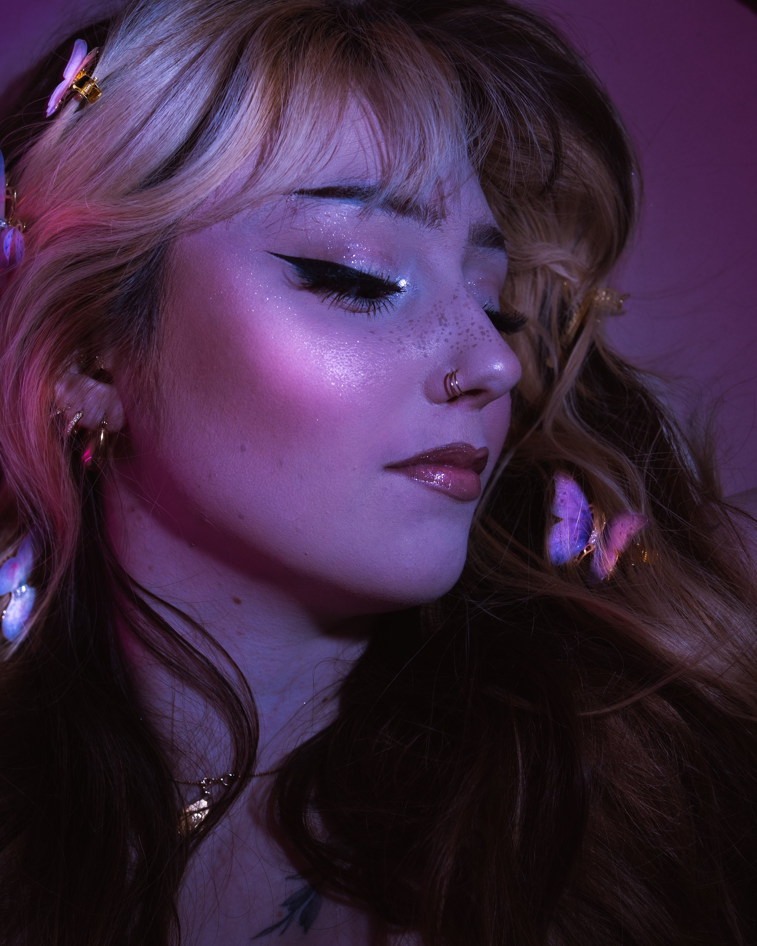 Une personne avec des cheveux bouclés, maquillage brillant et oreilles avec des accessoires, sous un éclairage violet. Shooting photo style edito.