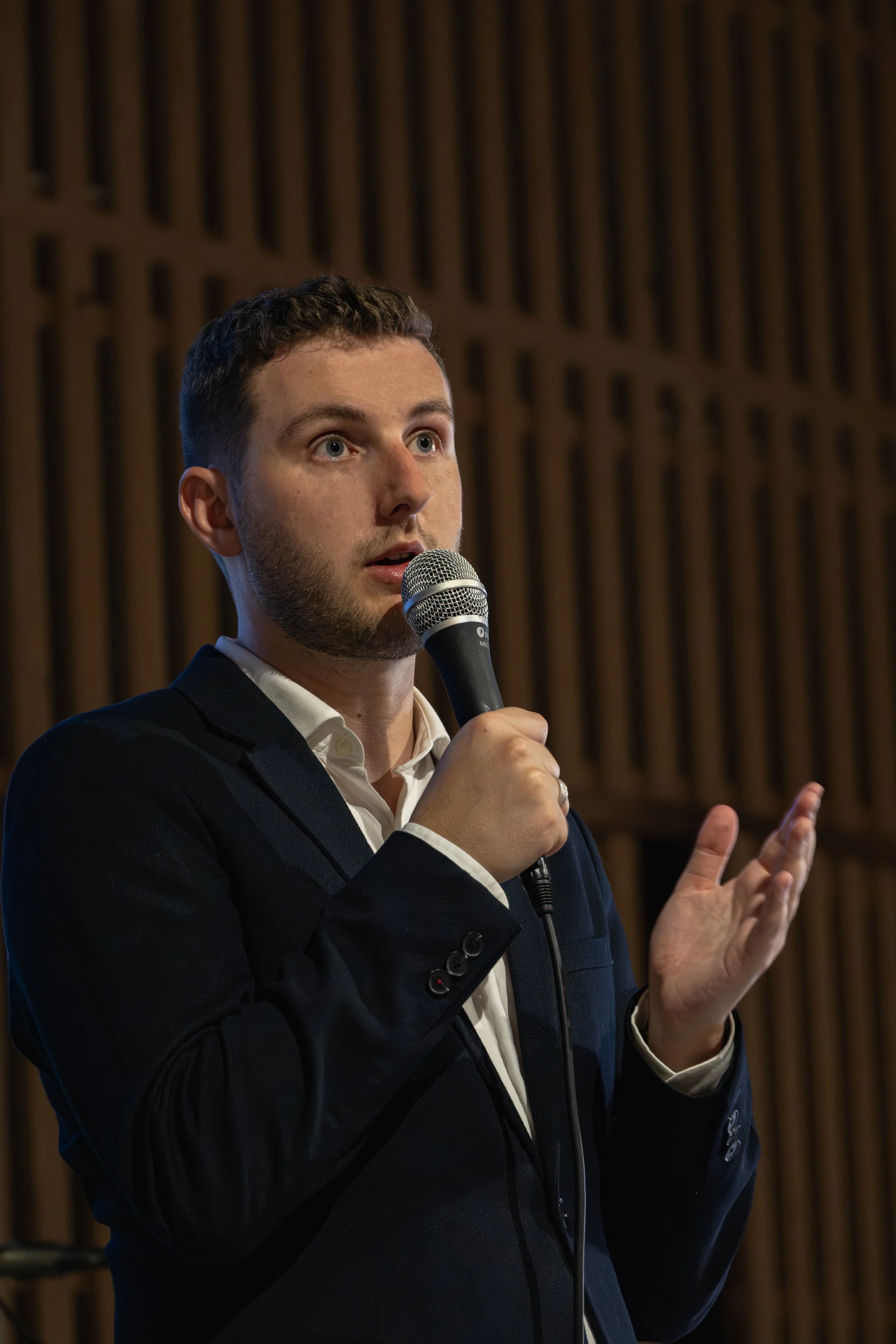 Un homme en costume noir parle dans un microphone lors d'une présentation ou d'un discours, avec un fond en bois.