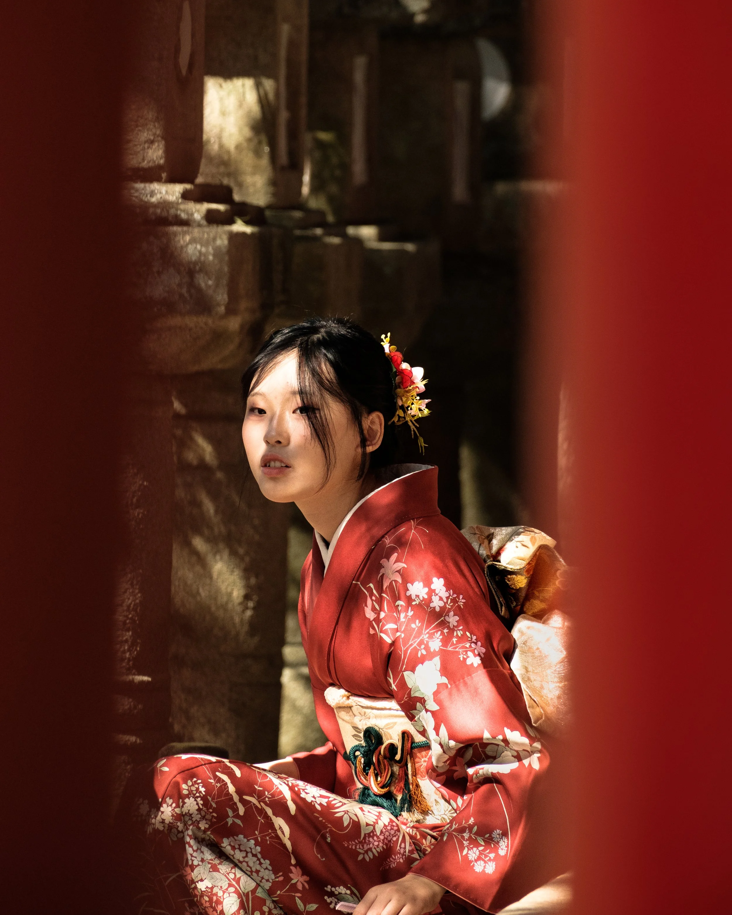 Jeune femme portant un kimono traditionnel japonais rouge avec des motifs floraux, coiffée d'une décoration florale dans les cheveux, assise dans un lieu sombre avec des éléments en pierre et en bois en arrière-plan.