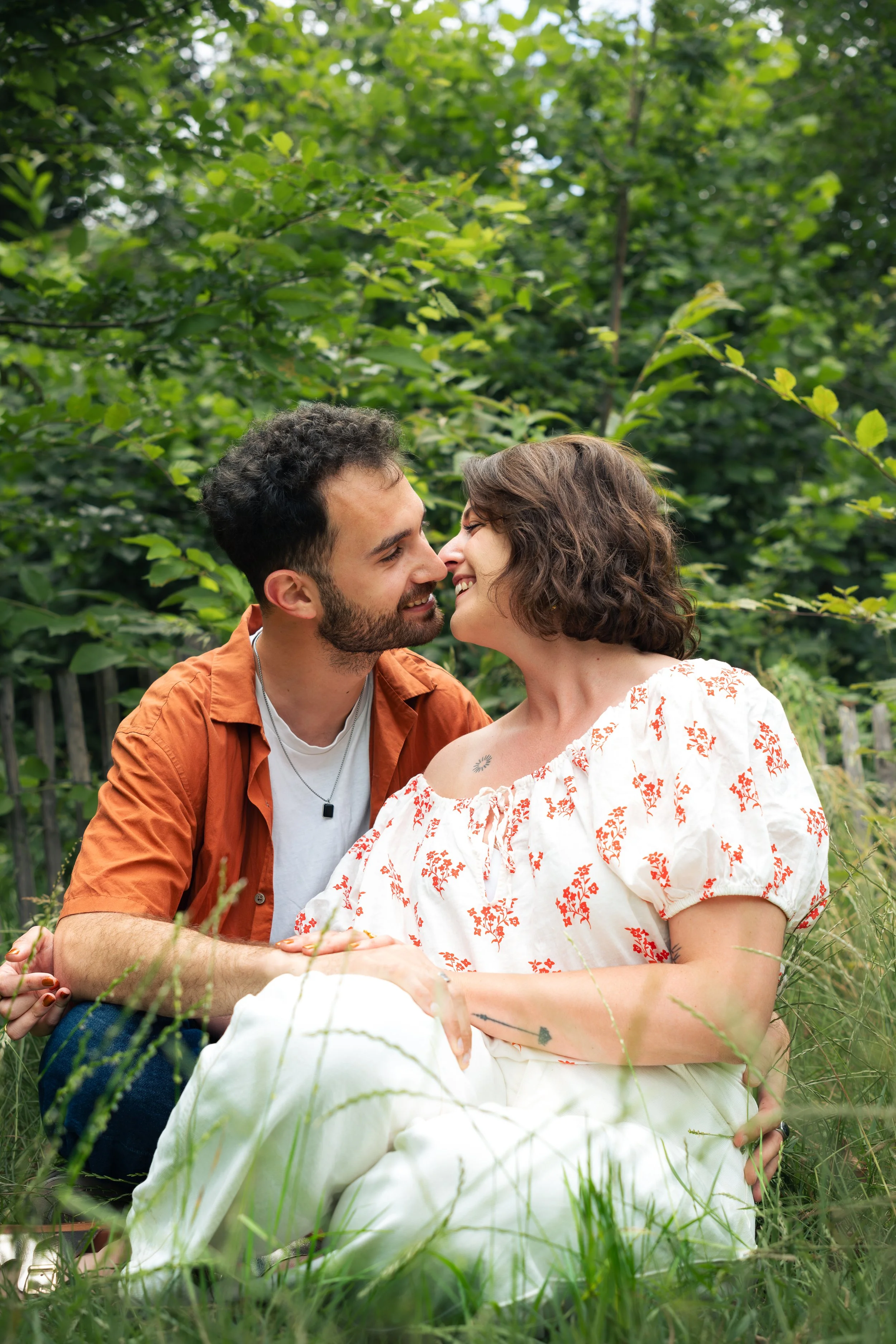 Un couple souriant face à face dans une nature verdoyante, assis sur l'herbe lors d'une séance photo.