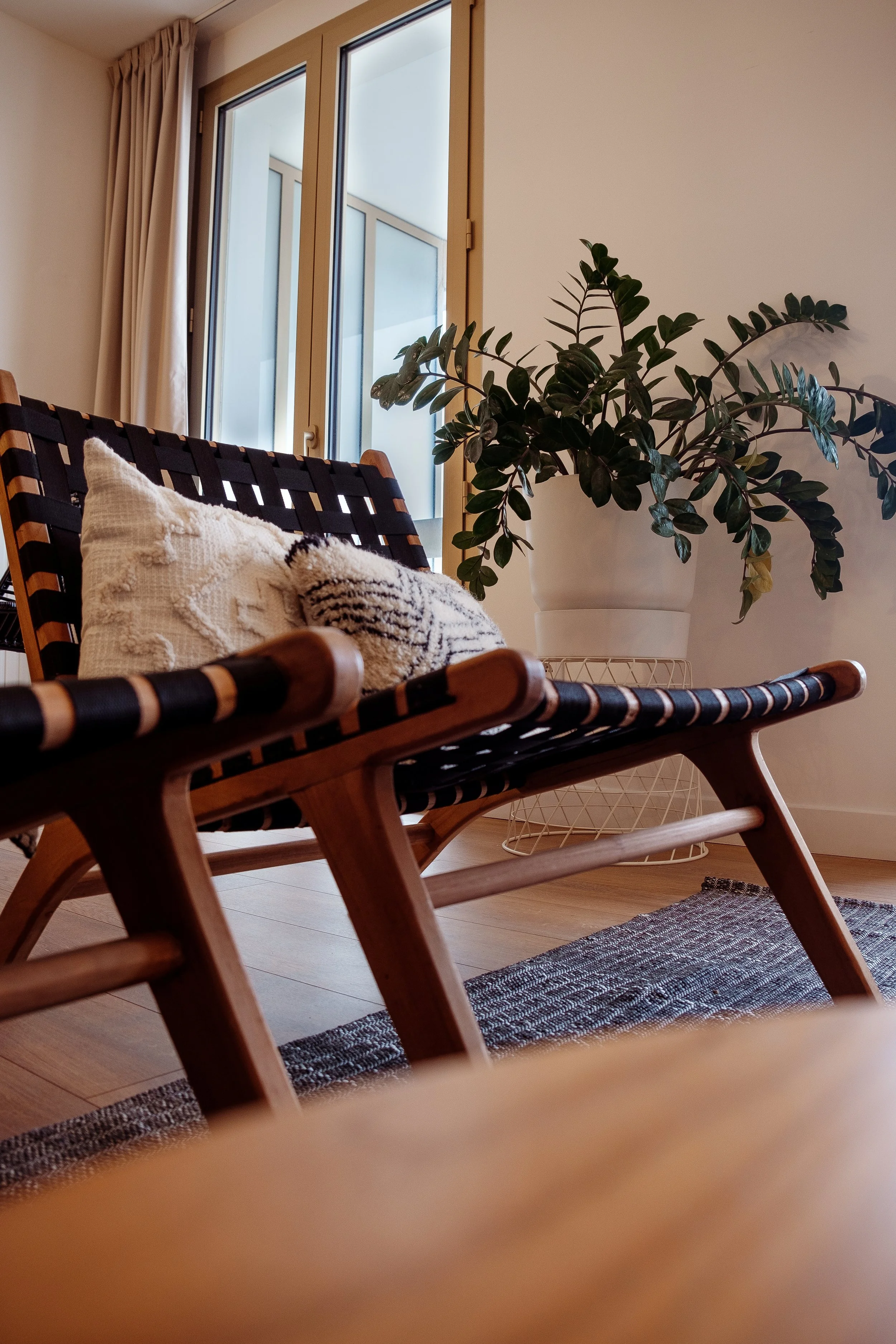 Une pièce intérieure avec un fauteuil en bois et tissu noir et beige, un coussin beige et noir, une plante verte dans un pot blanc, un mur blanc, une fenêtre avec des rideaux beige, un tapis gris et un sol en bois.
