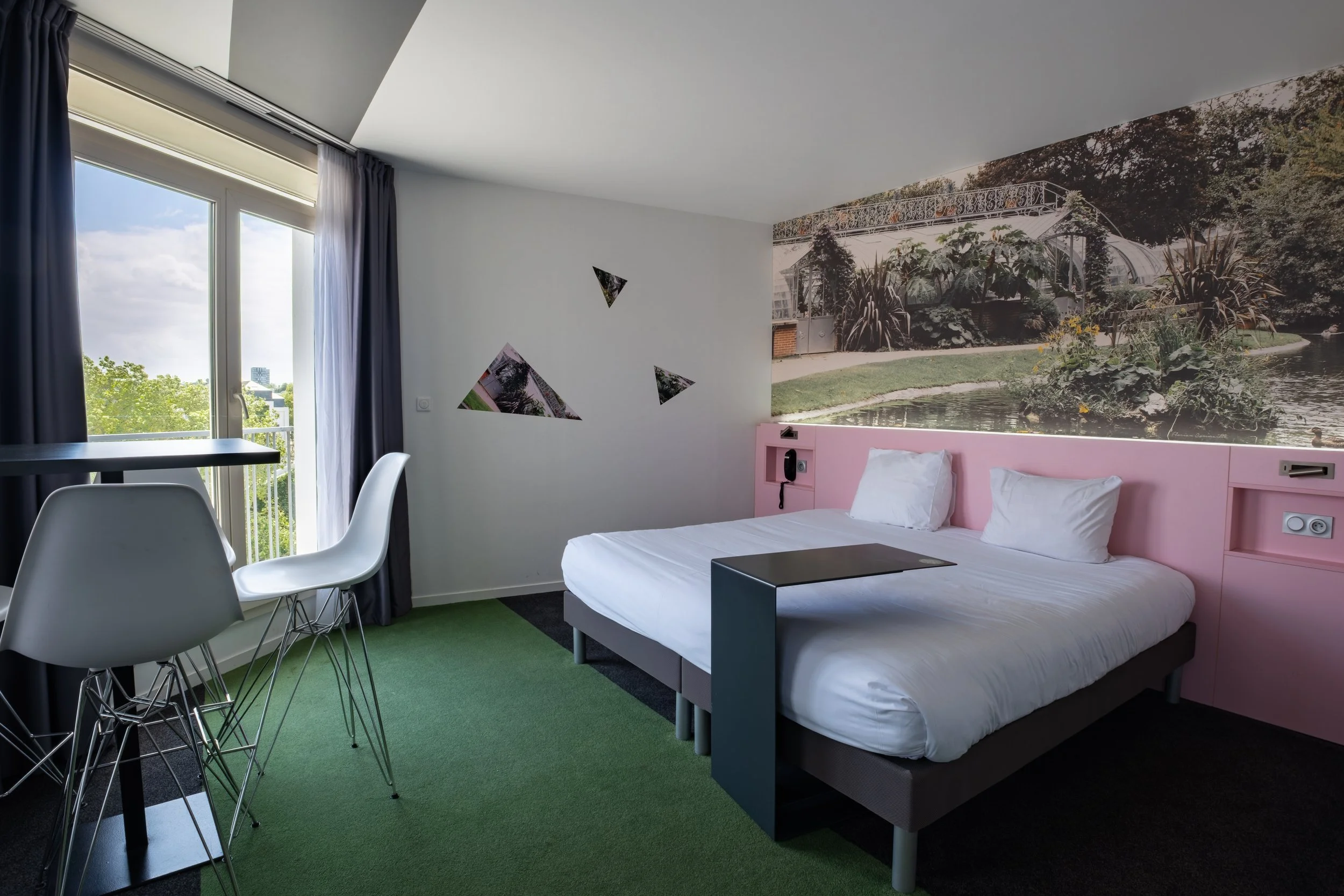 Chambre d'hôtel avec un lit double, un mur avec une fresque représentant un jardin avec une serre, un balcon avec vue sur des arbres. Fenêtre avec rideaux gris foncé. Un coin repas avec une table noire et quatre chaises blanches. Parois blanches et t