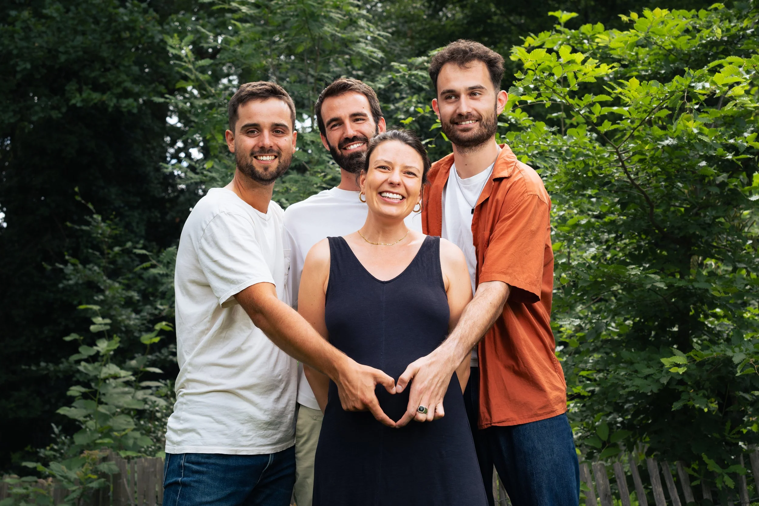 Cinq personnes souriantes dans une forêt, se tenant par les mains et formant un cœur avec leurs bras. Photo d'un groupe d'ami pour annoncer une grossesse, et un faire part de naissance.
