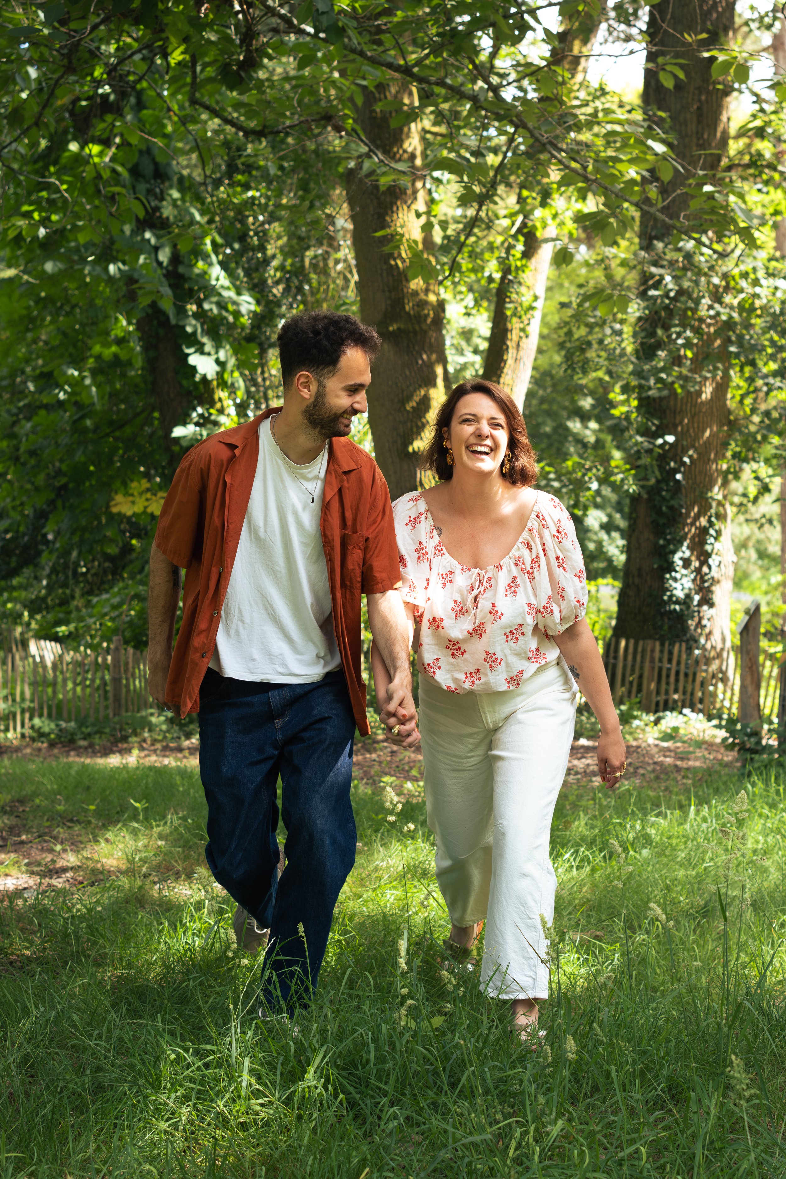 Un couple joyeux marchant main dans la main dans un parc verdoyant avec des arbres en arrière-plan, pour un shooting photo.