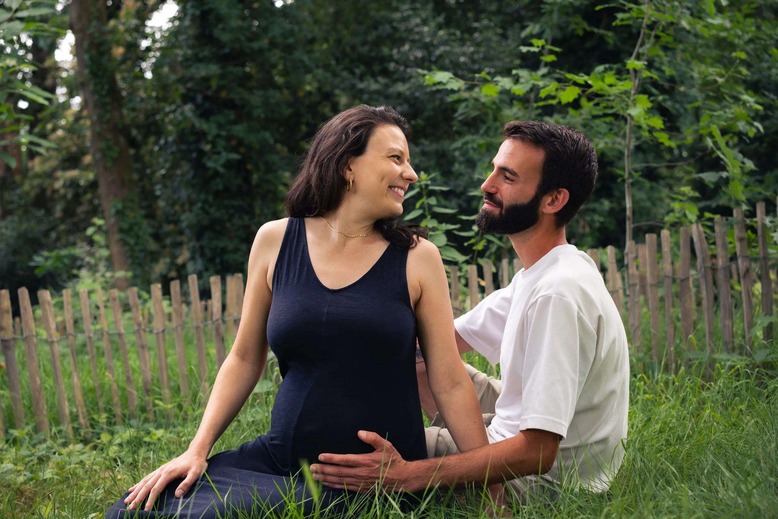 Un couple partage un moment intime dans un jardin avec des arbres verts, la femme enceinte assise sur la pelouse et l'homme à genoux, tous deux souriants et regardant dans les yeux.