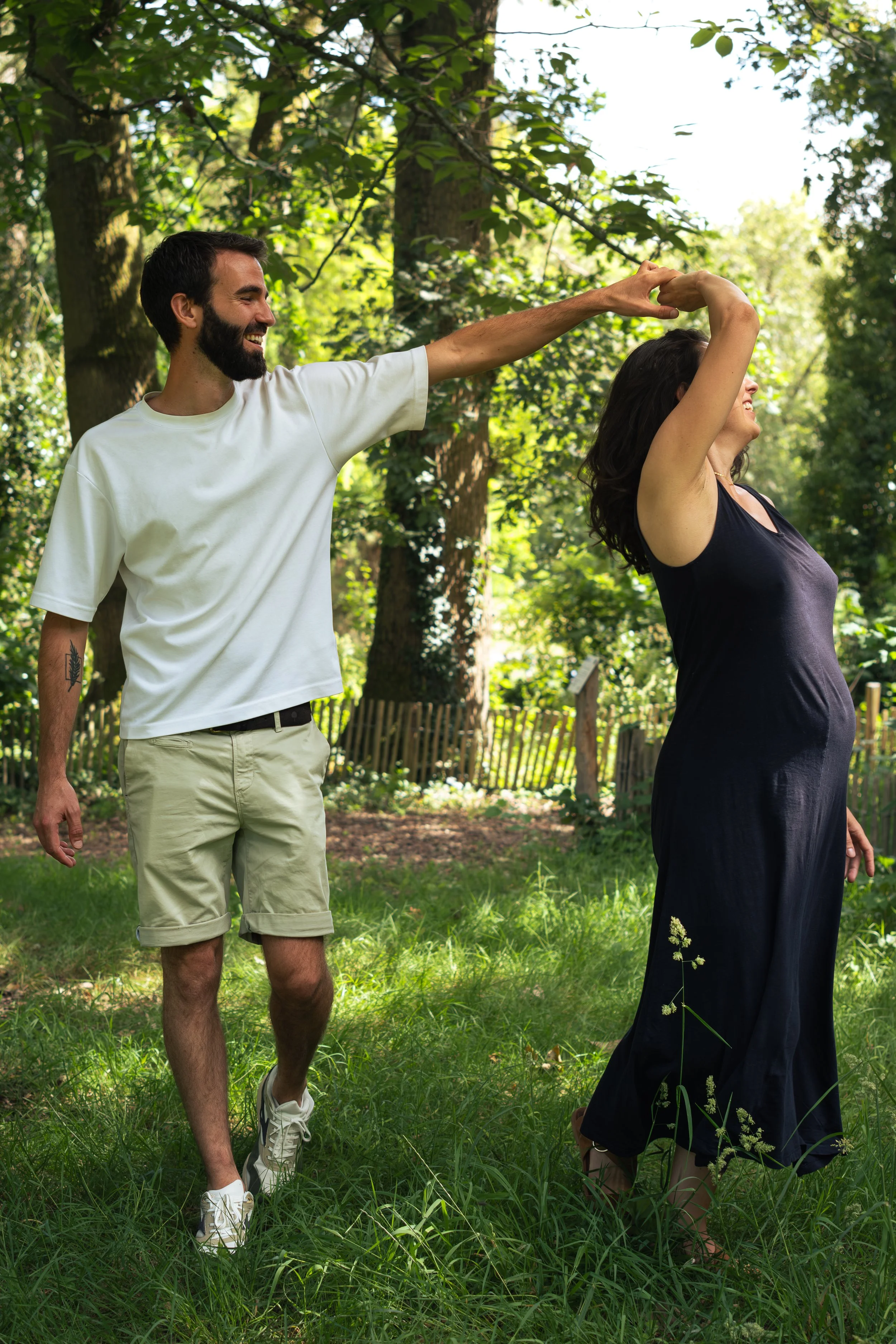 Un homme et une femme dans un parc, l'homme tire la femme par la main en dansant, tous deux souriants, entourés d'arbres et d'herbe verte.