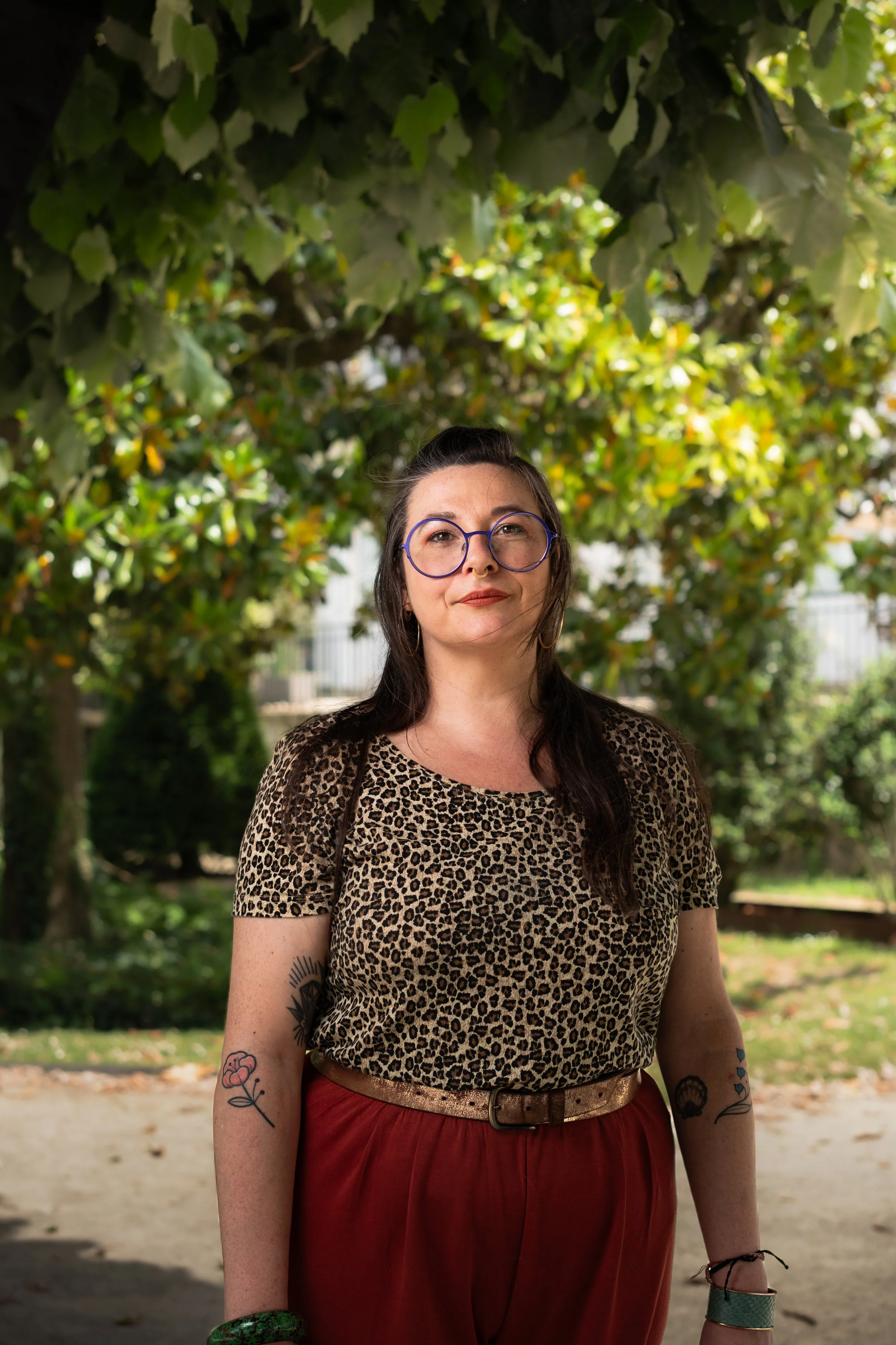 Jeune femme debout dans un parc, portant une chemise léopard, des lunettes rondes, des tatouages sur les bras, et des bracelets, avec un fond de feuillage verdoyant. C'est un portrait casual corporate