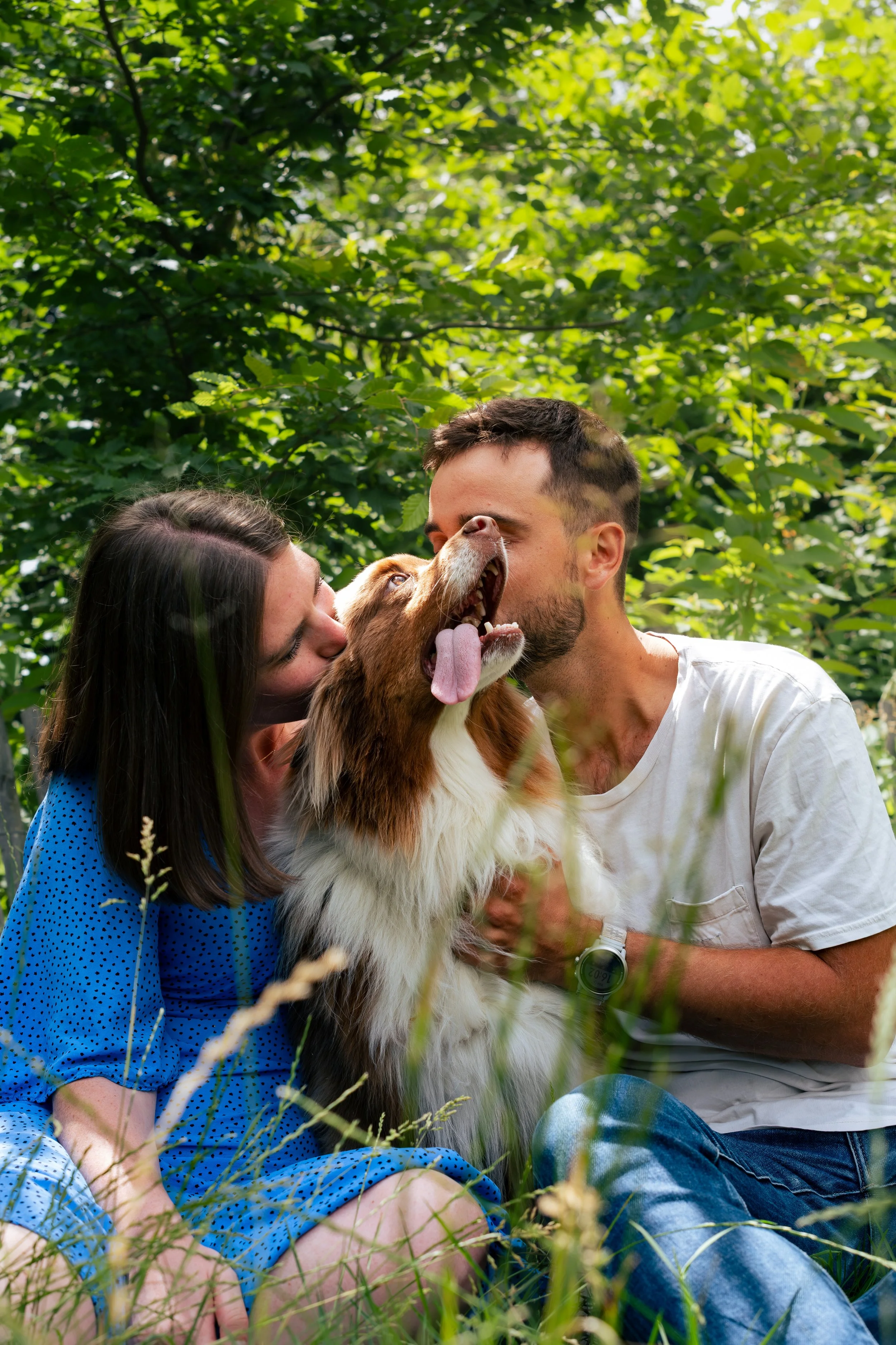 Un couple et un chien dans un parc verdoyant. La femme porte une robe bleue à pois et l'homme un t-shirt blanc. Ils semblent s'amuser avec le chien, qui a la langue sortie et semble heureux.