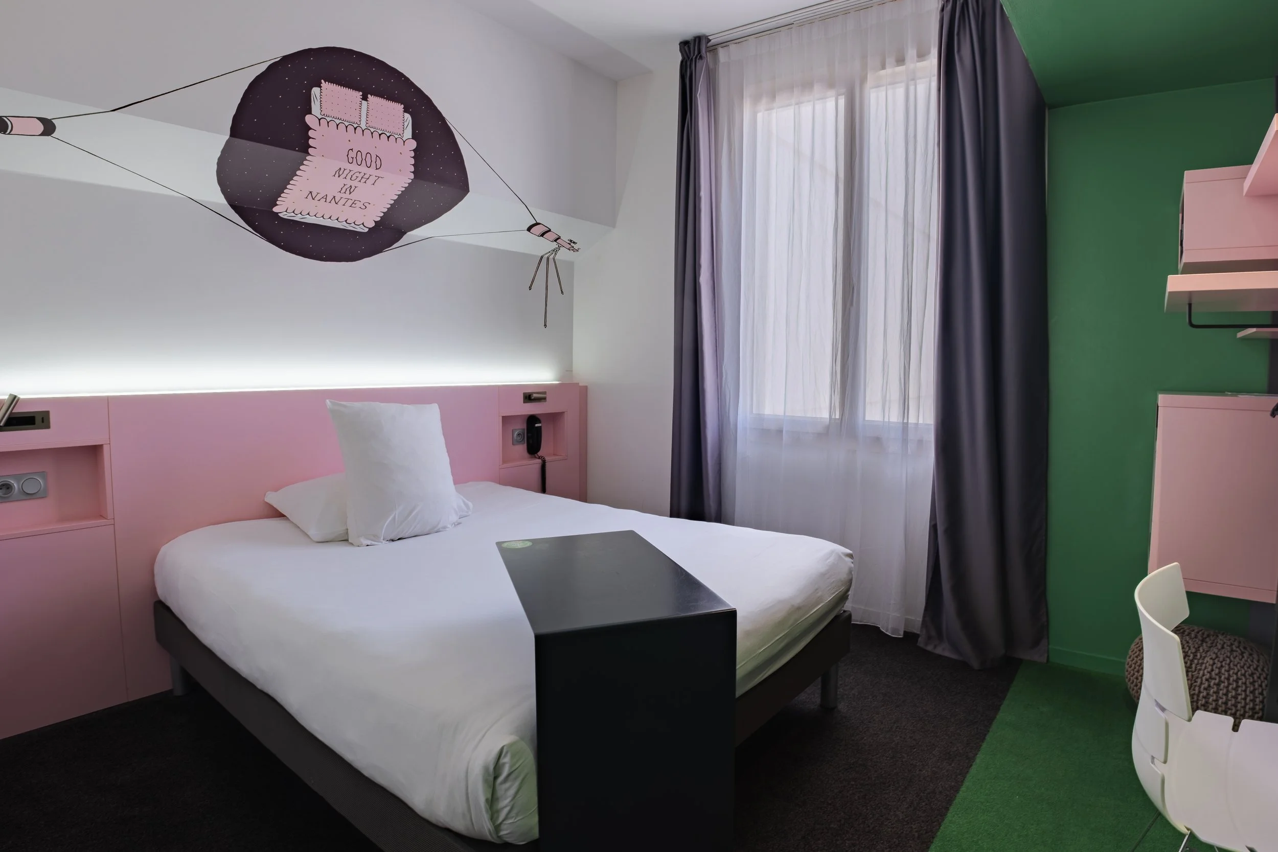 Chambre d'hôtel moderne avec un lit simple, une tête de lit rose, un bureau noir, un mur vert et une fenêtre avec rideaux gris et blancs. Un mur au plafond orné d'une illustration d'une lune, d'une étoile et d'une boussole, avec le texte "Good night 