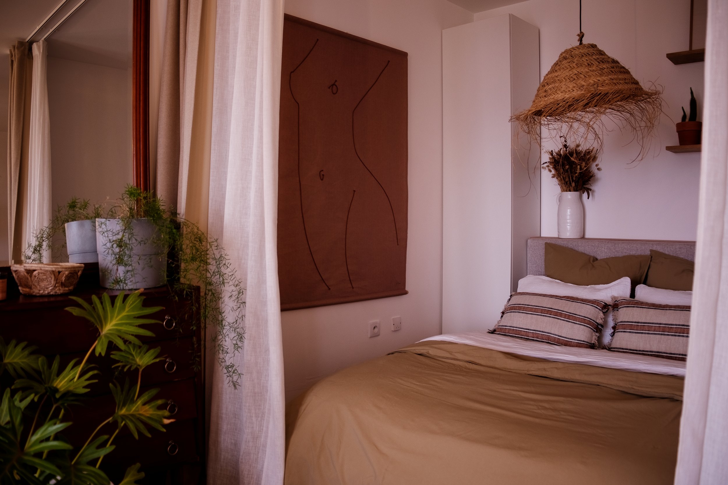Une chambre avec un lit fait, des oreillers, une plante verte, une commode en bois avec des objets décoratifs, un tableau abstrait en bois, une lampe en osier suspendue, et des étagères avec des plantes en pot.