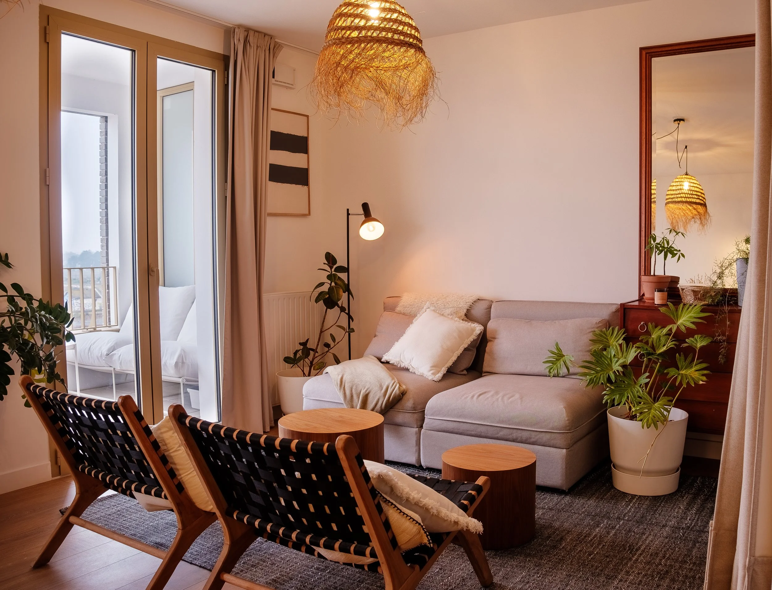 Un salon avec un canapé gris, plusieurs plantes en pots, deux chaises en bois et un tabouret. Il y a une grande fenêtre et un miroir mural qui reflète un lampadaire et un lustre suspendu.