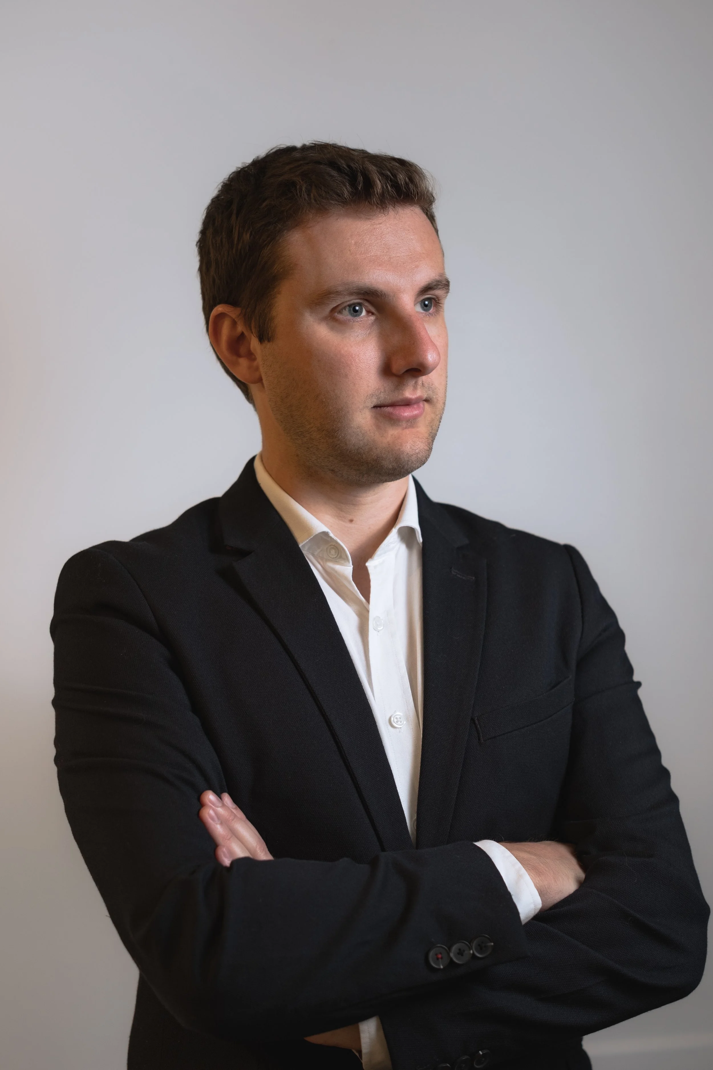 Homme en costume noir croisé les bras, regardant vers la droite avec une expression sérieuse, portant une chemise blanche et un fond gris clair