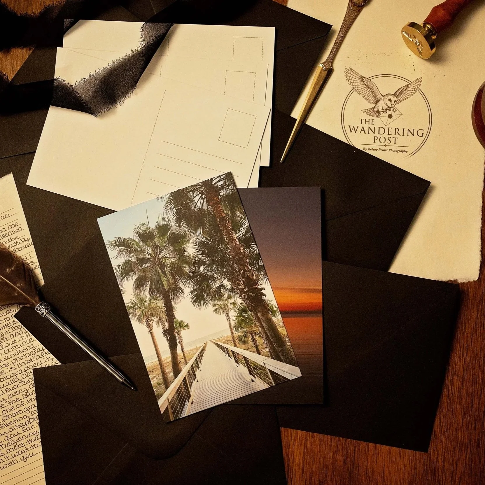 Photograph of a desk with tropical vacation photos, black envelopes, a silver pen, a gold and silver letter opener, and a logo reading "The Wandering Post" with an owl, on a wooden surface.