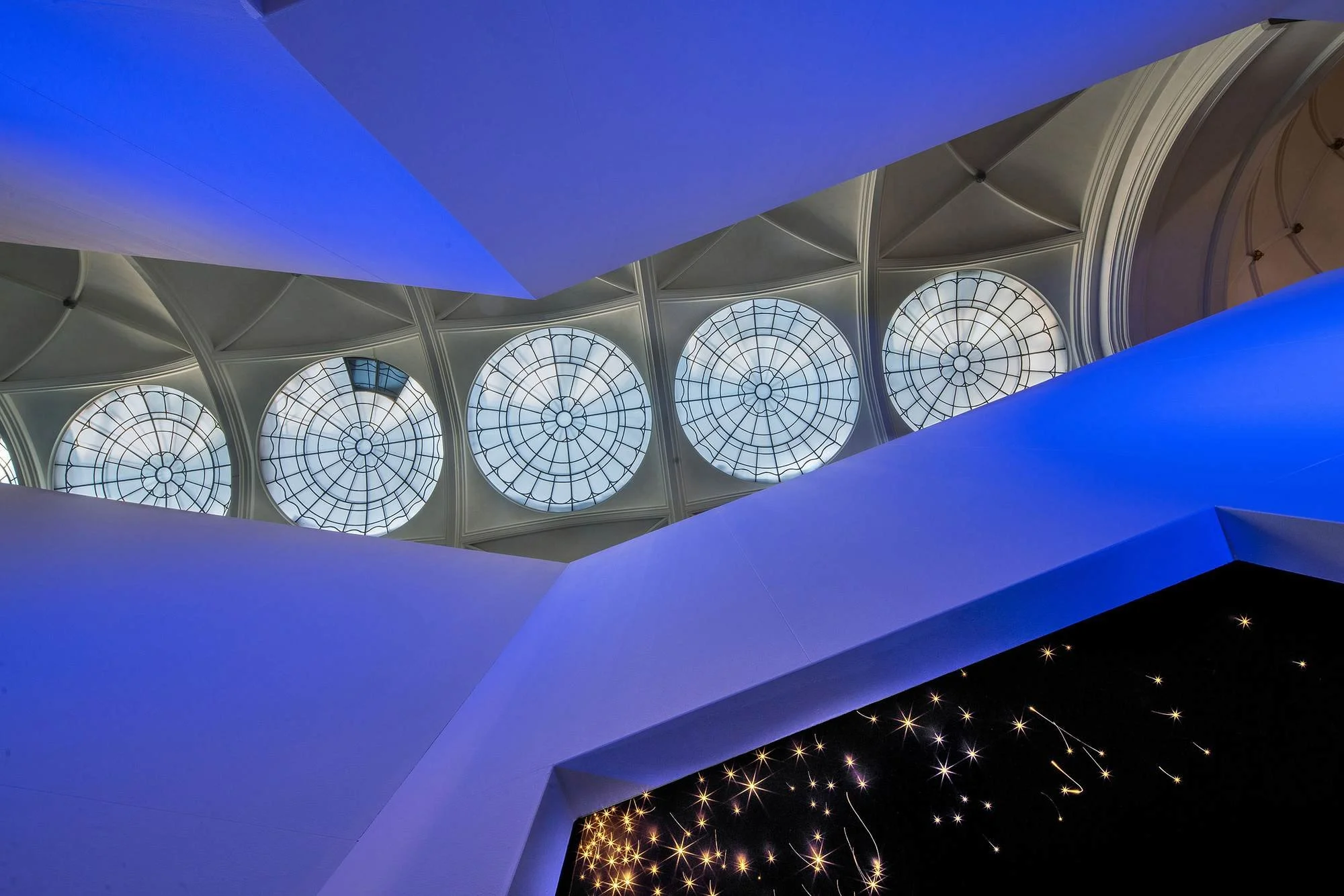 The image shows the interior of a museum with large circular stained glass windows on the ceiling, illuminated by blue lighting, with architectural white walls and a dark background with star-like lights.