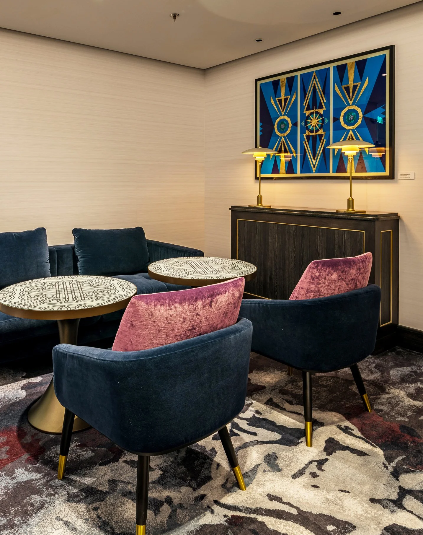 A stylish lounge area with velvet chairs in blue and pink, round tables with intricate black and white patterns, a black and gold sideboard, two gold lamps, and a colorful, geometric abstract gilded glass artwork on the wall.