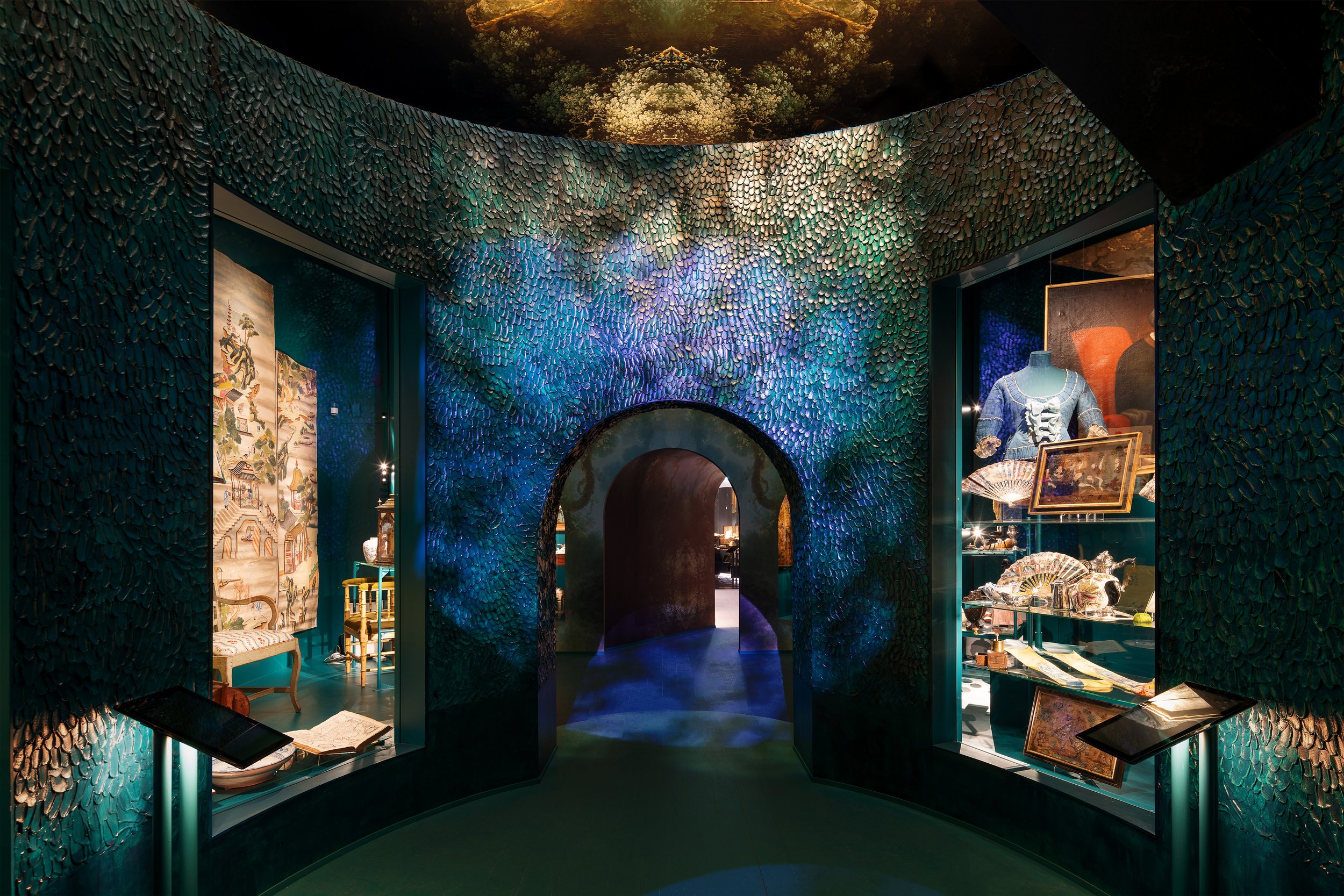 Interior of a museum or exhibit space featuring vibrant, textured walls made of overlapping plaster leaves in shades of green, blue, and purple. 