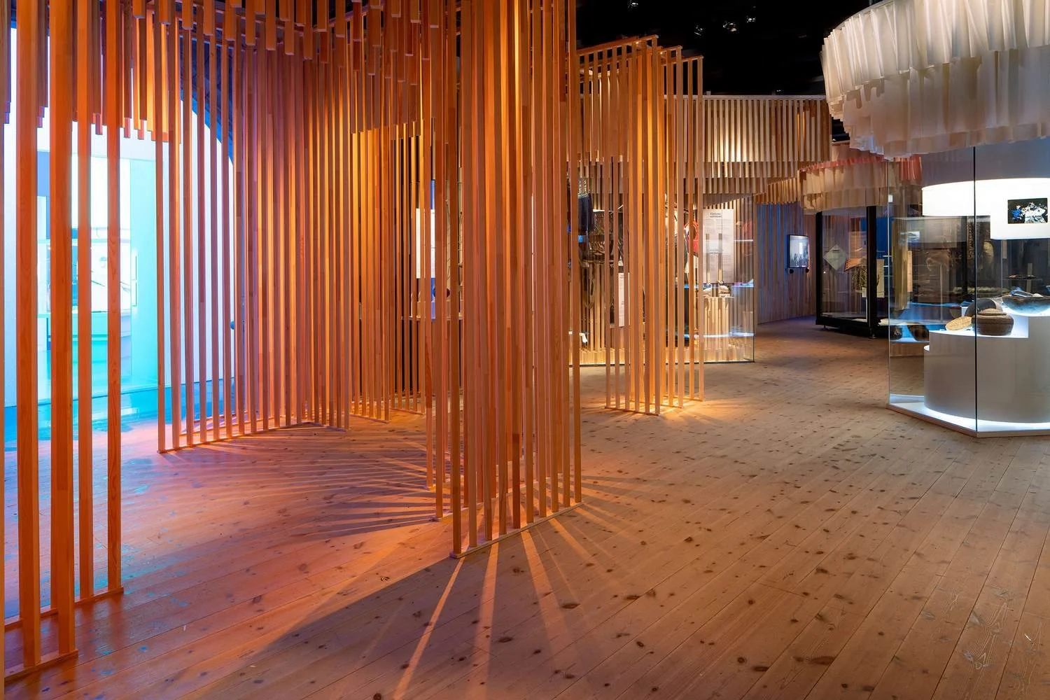 Interior of a museum with wooden vertical slat partitions, glass display cases, and artifacts on white stands, illuminated with warm lighting.