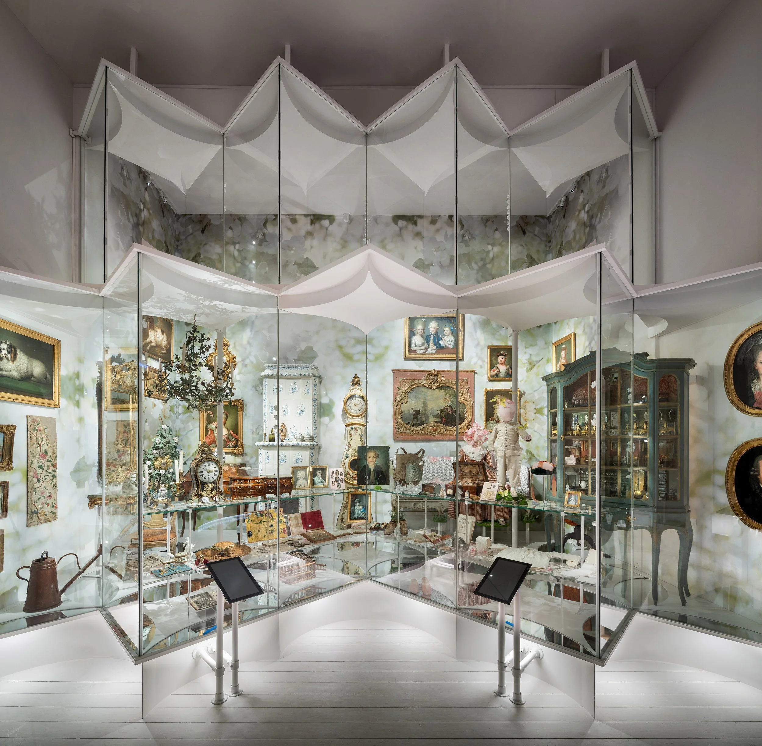 An interior view of a museum or gallery with a mirrored display case filled with antique items, paintings on the walls, and decorative furniture.