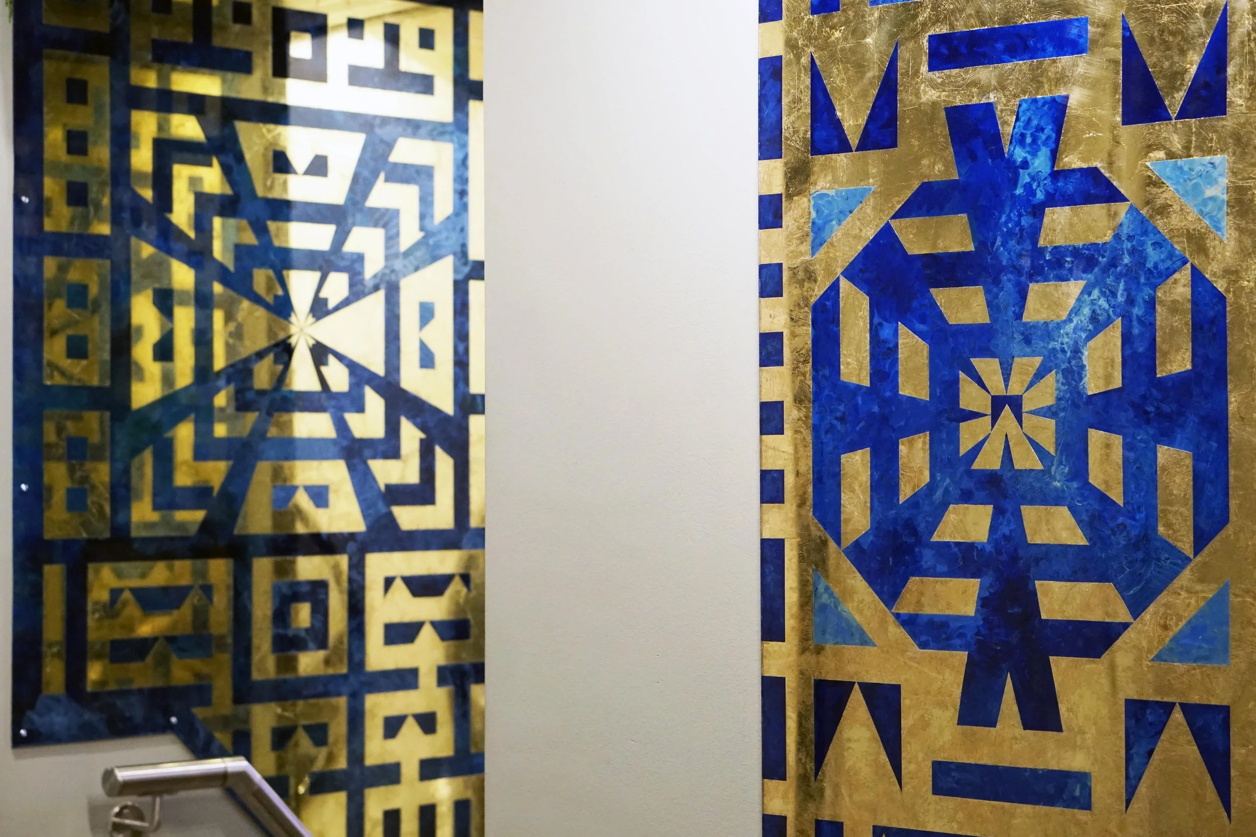 Two pieces of artwork with geometric patterns, one on the left with yellow and blue shapes, one on the right with blue and gold shapes, separated by a plain white wall.