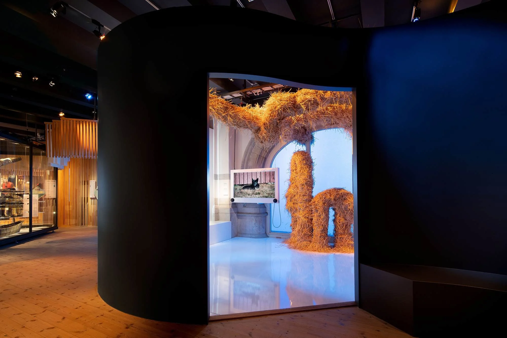 An art installation featuring a large, straw-like sculpture resembling a creature or abstract form, situated in a museum with natural light and a window in the background.