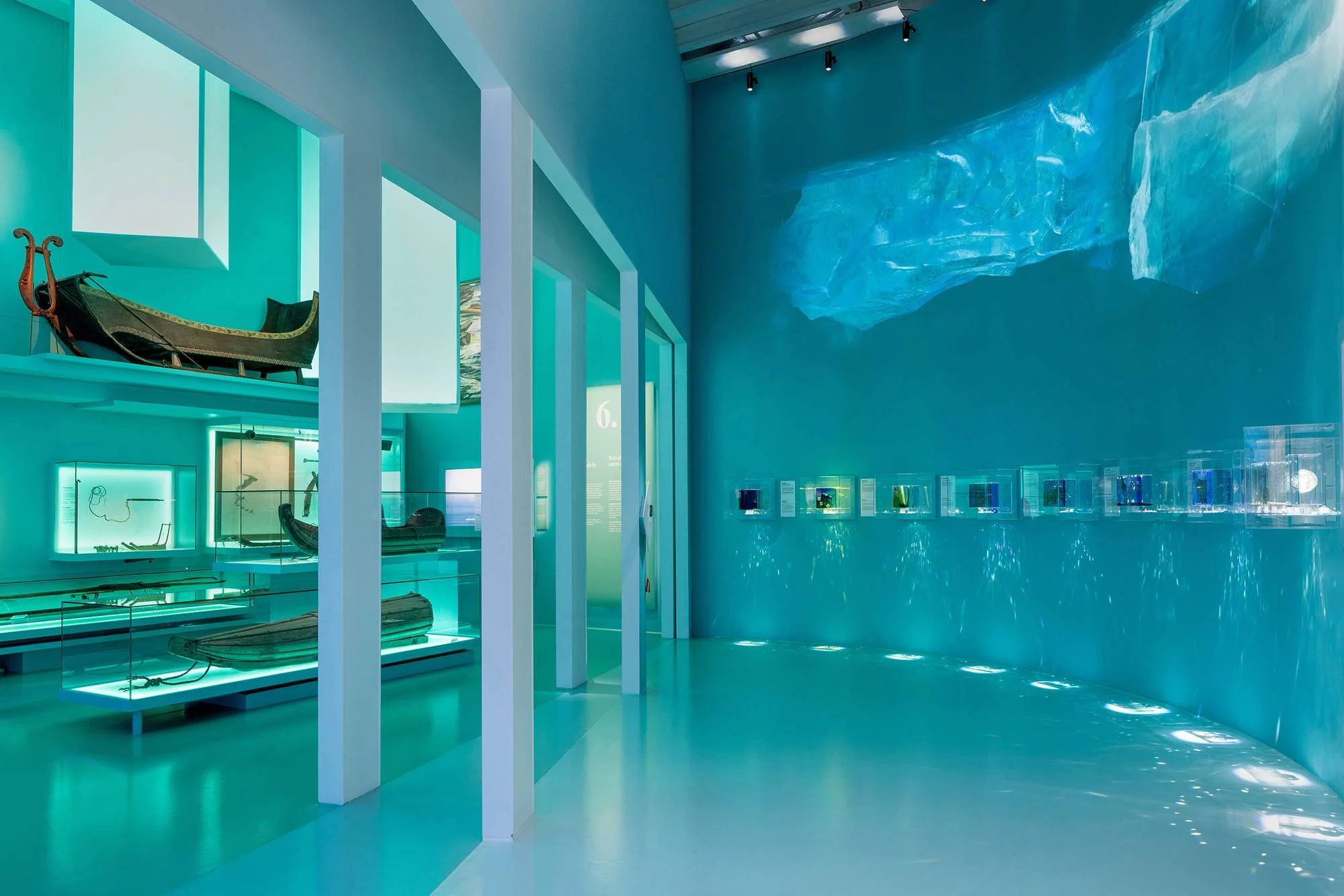A modern art gallery interior with turquoise walls and glossy flooring. Exhibited artifacts include a vintage sleigh on a raised platform, glass display cases with various objects, and a large abstract artwork on the wall. The ceiling has track light