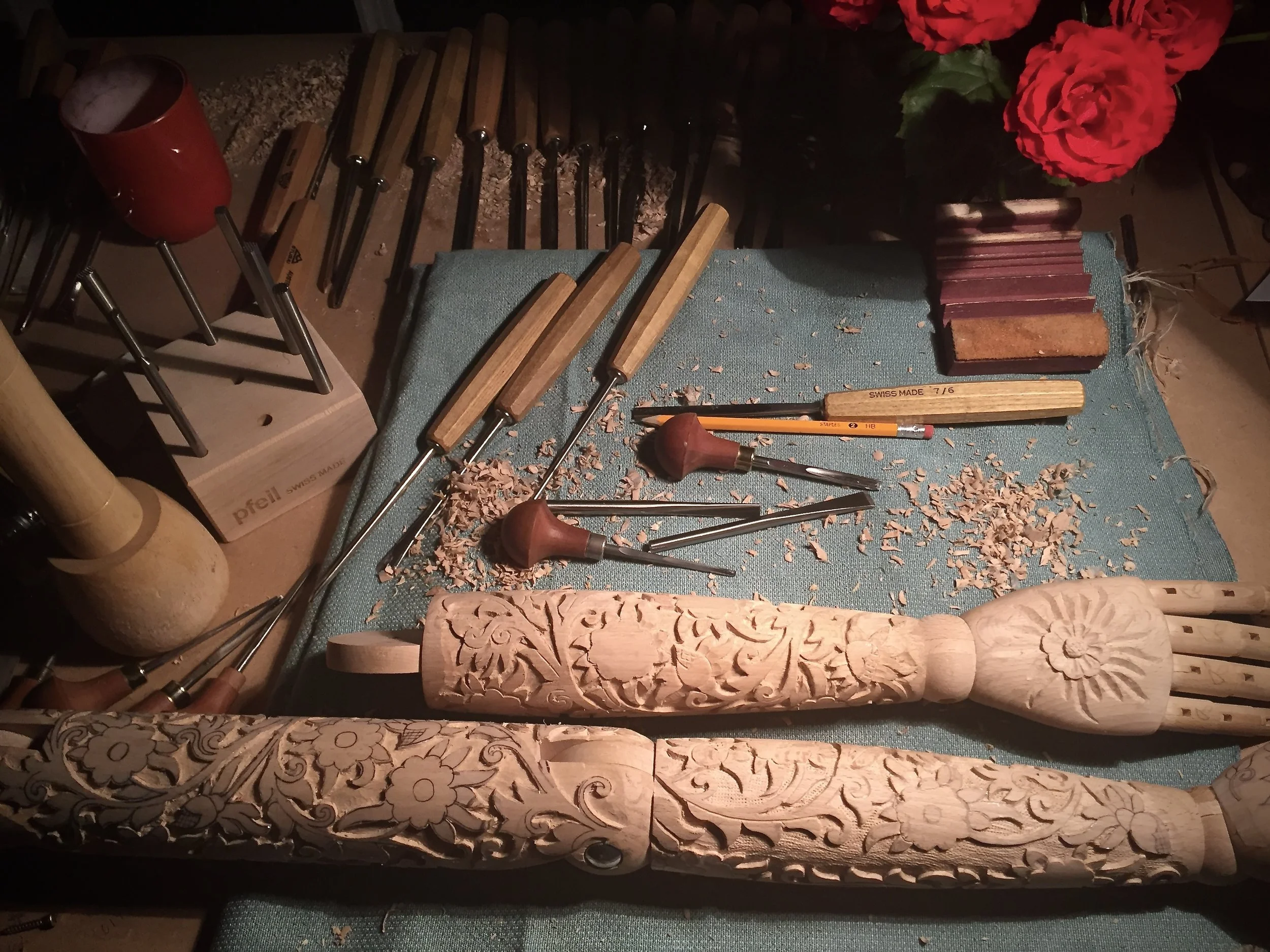 Carving tools and a wooden mannequin arms with floral and swirl patterns, carving shavings, on a worktable with a textured blue cloth, red flowers in a vase in the background.