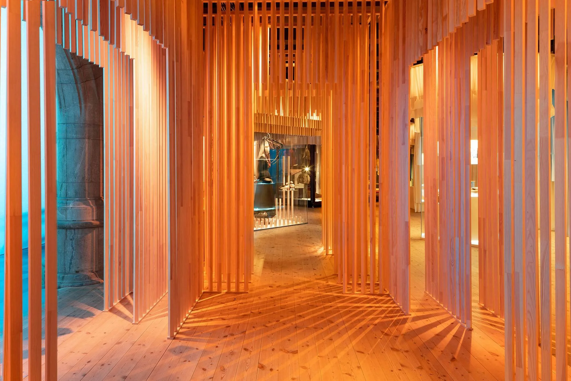 Interior view of a building with wooden vertical slats creating a corridor-like space, with warm lighting illuminating the wooden floor and walls.