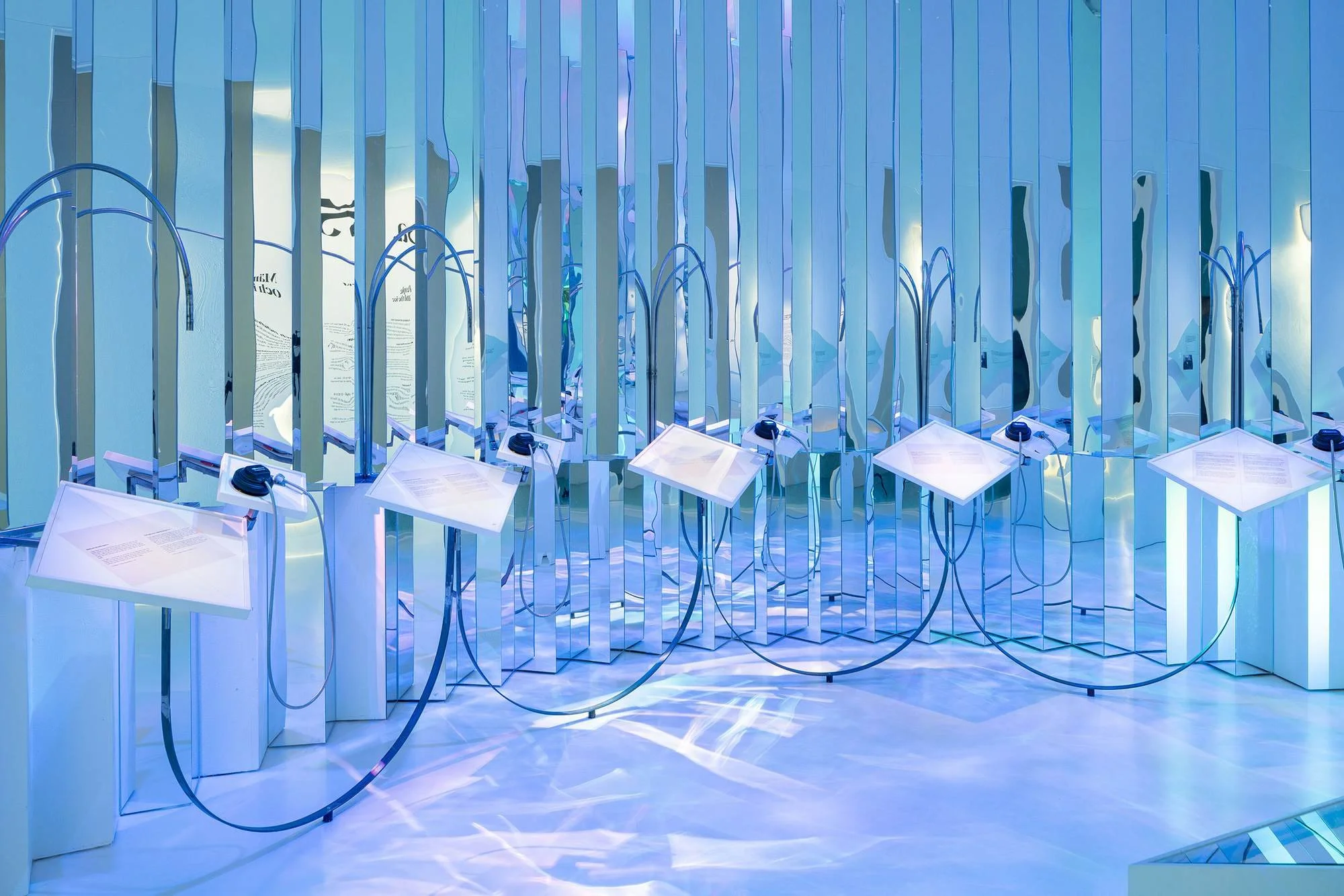 A modern exhibition space with reflective, vertical, blue-tinted panels and display stands with informational plaques, connected by black cords, all illuminated by blue lighting.