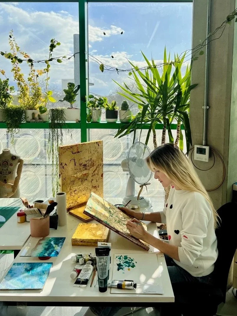 The artist painting at a desk with various art supplies, sitting in a bright studio with plants and large windows showing a blue sky.