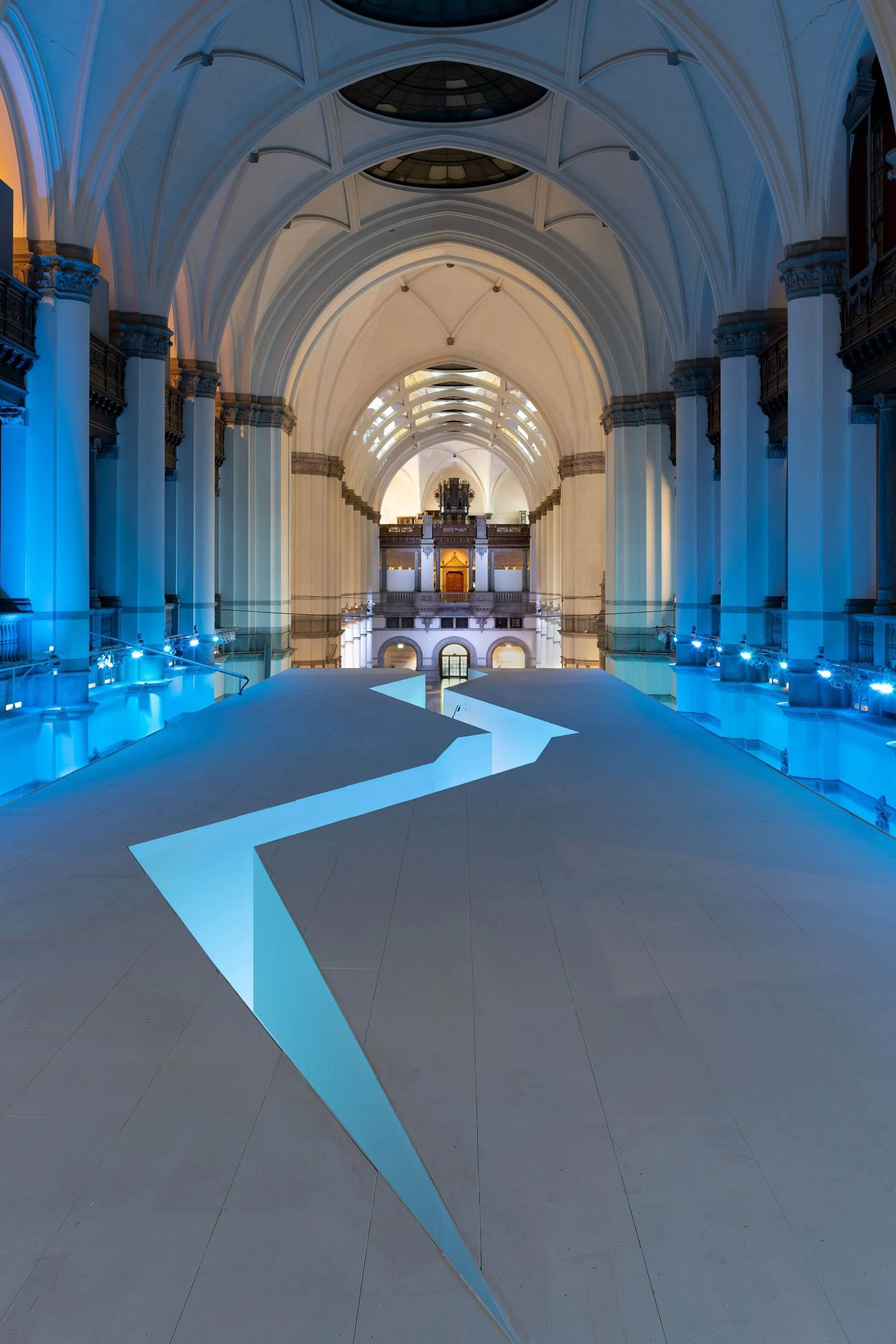 Interior of a grand, historic building with tall arches, columns, and a high ceiling, illuminated with blue lighting, Nordiska Museet.