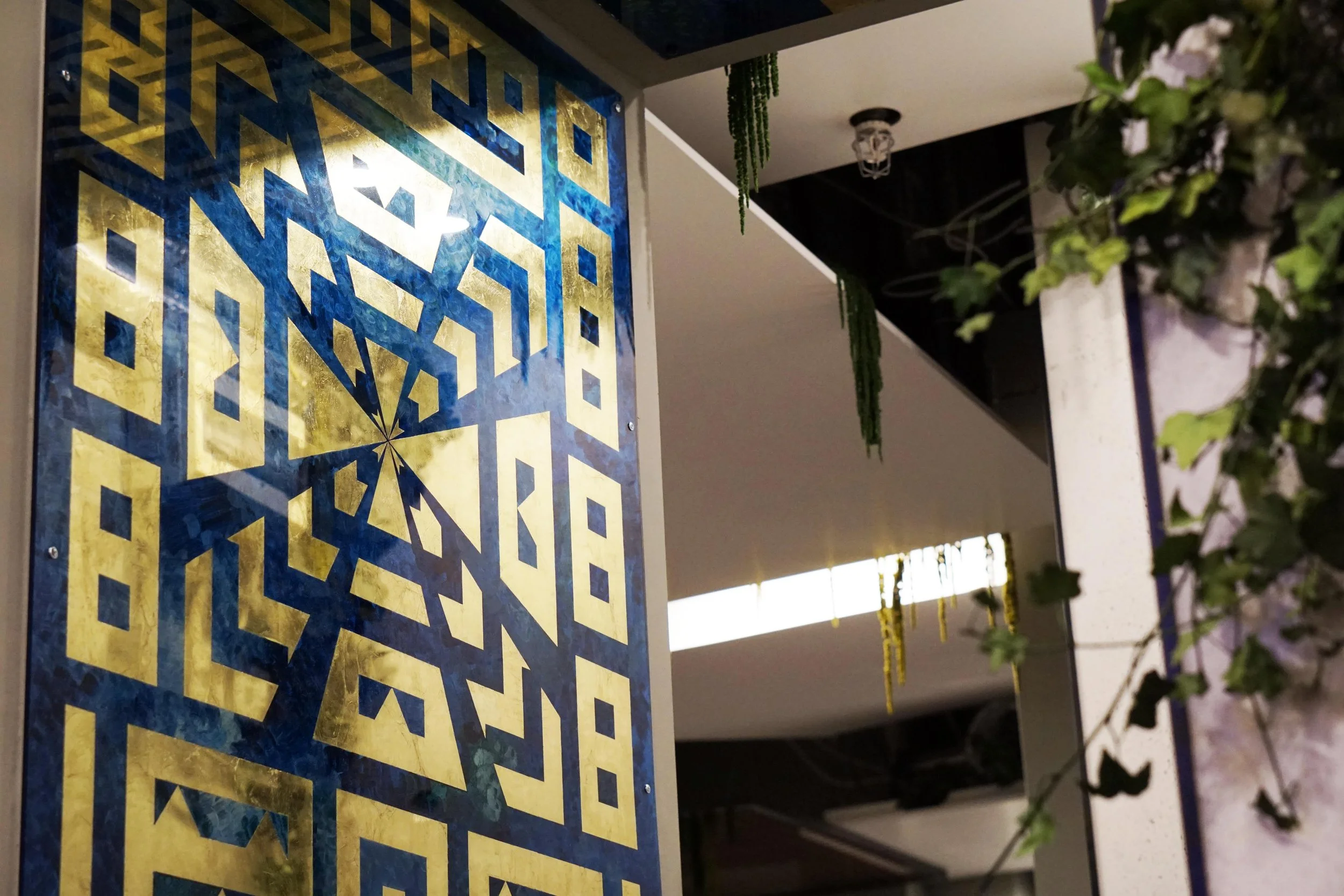 A decorative wall panel with geometric patterns in blue and gold, hanging in an indoor space with hanging plants and greenery, and a ceiling with lighting fixtures.