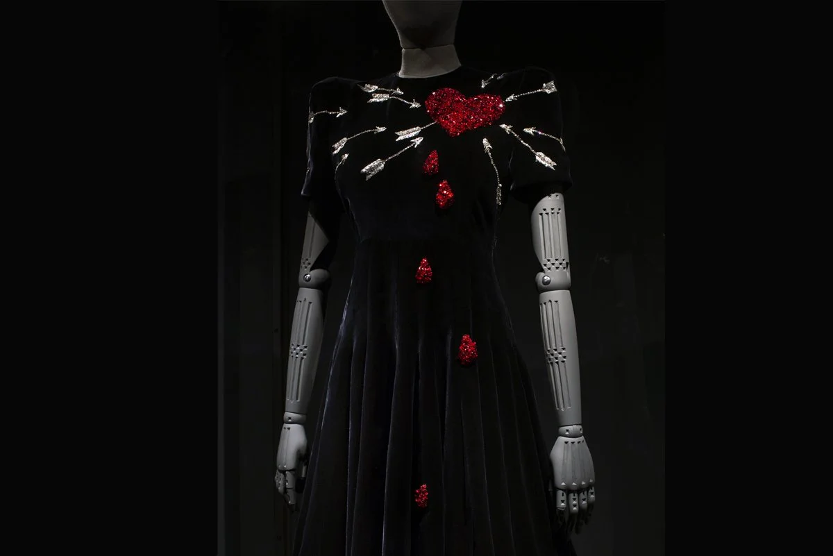 A black dress with red heart and drop-shaped embellishments on the chest and lower front, displayed on a mannequin with hand-carved arms against a dark background.