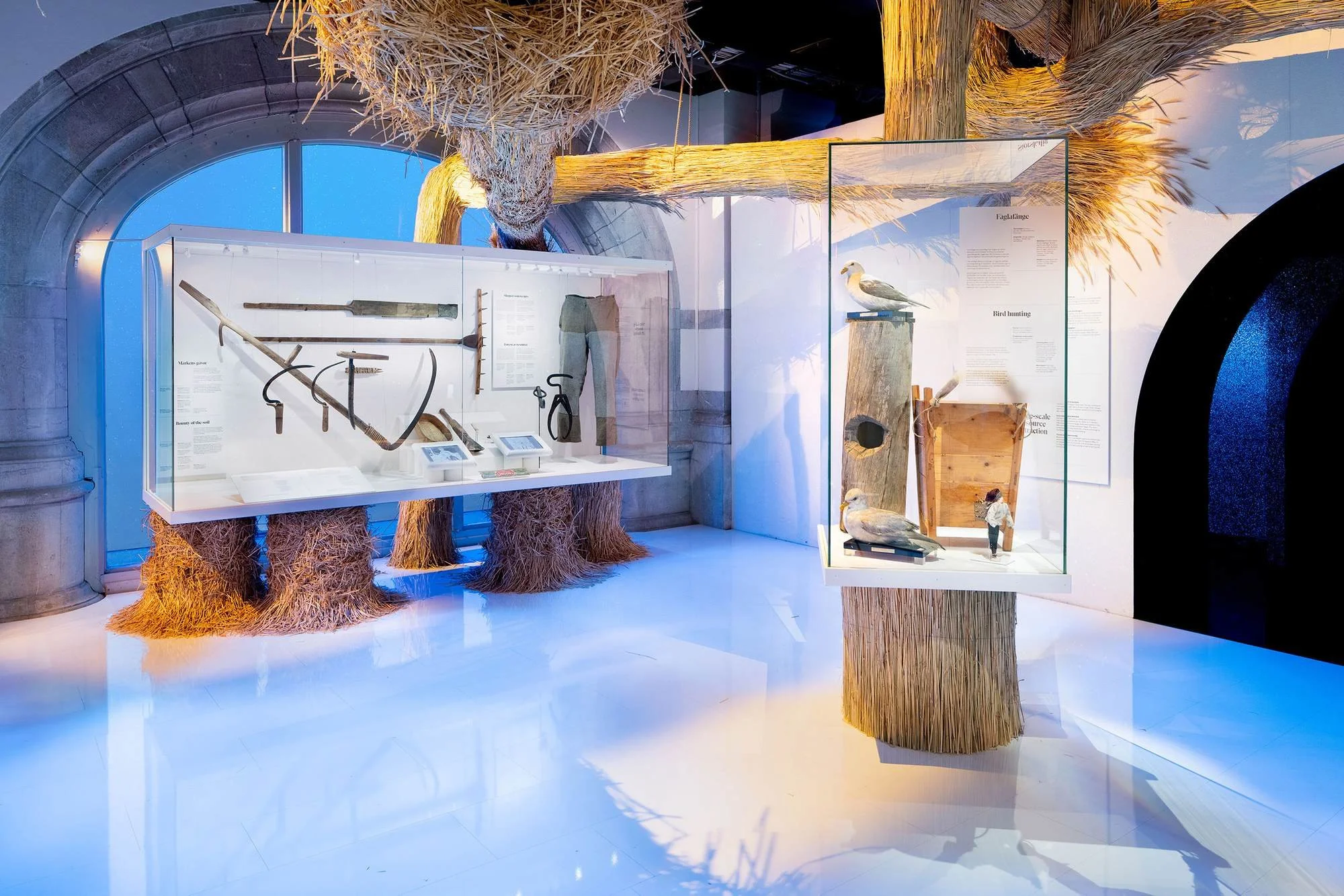 Museum exhibit featuring vintage farming tools, a model bird, and sculpted straw decorations with a large arched window in the background.