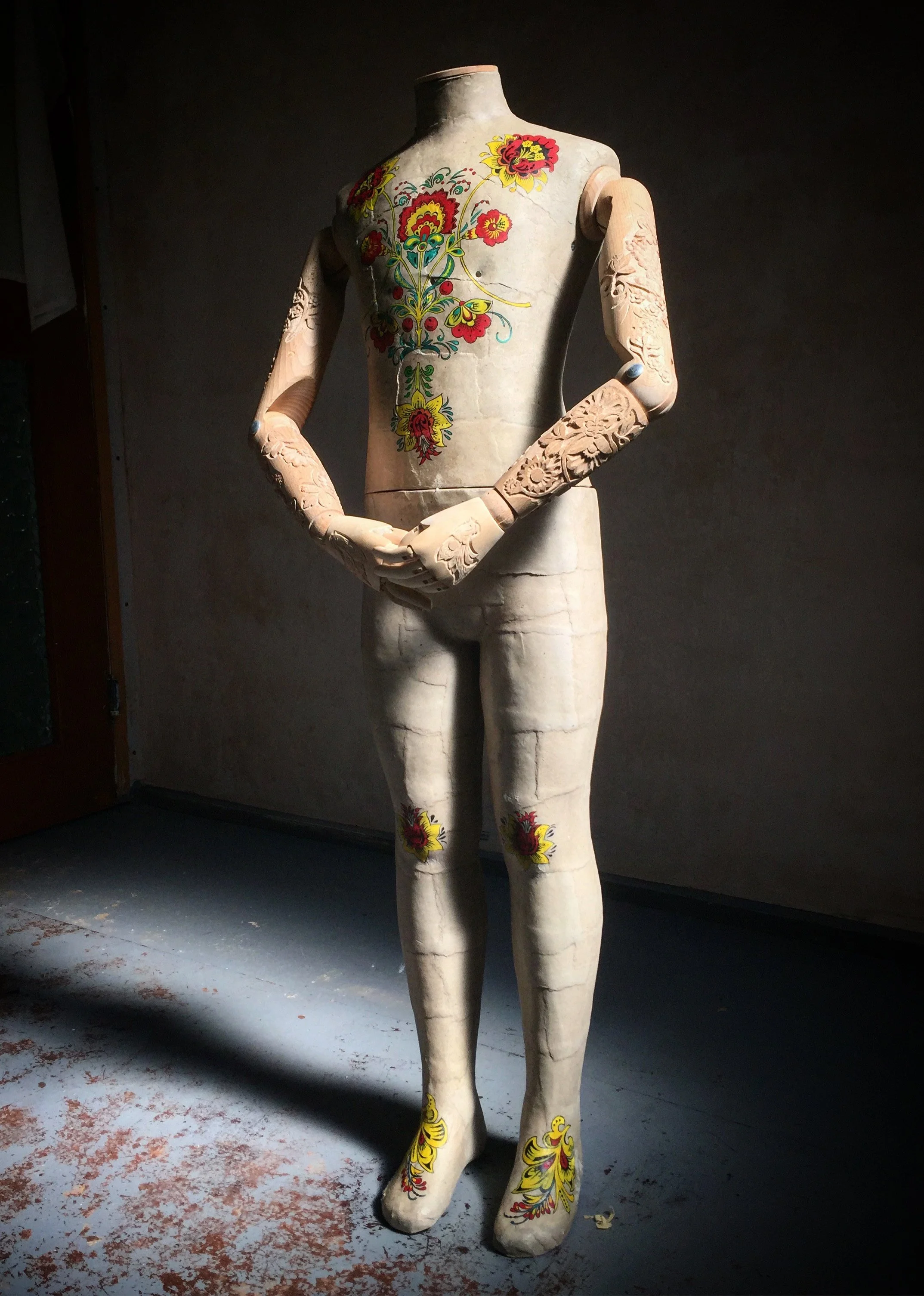 A mannequin with floral hand-painted designs on its torso and wood-carved arms, positioned in a dimly lit room with a gray floor and dark background.