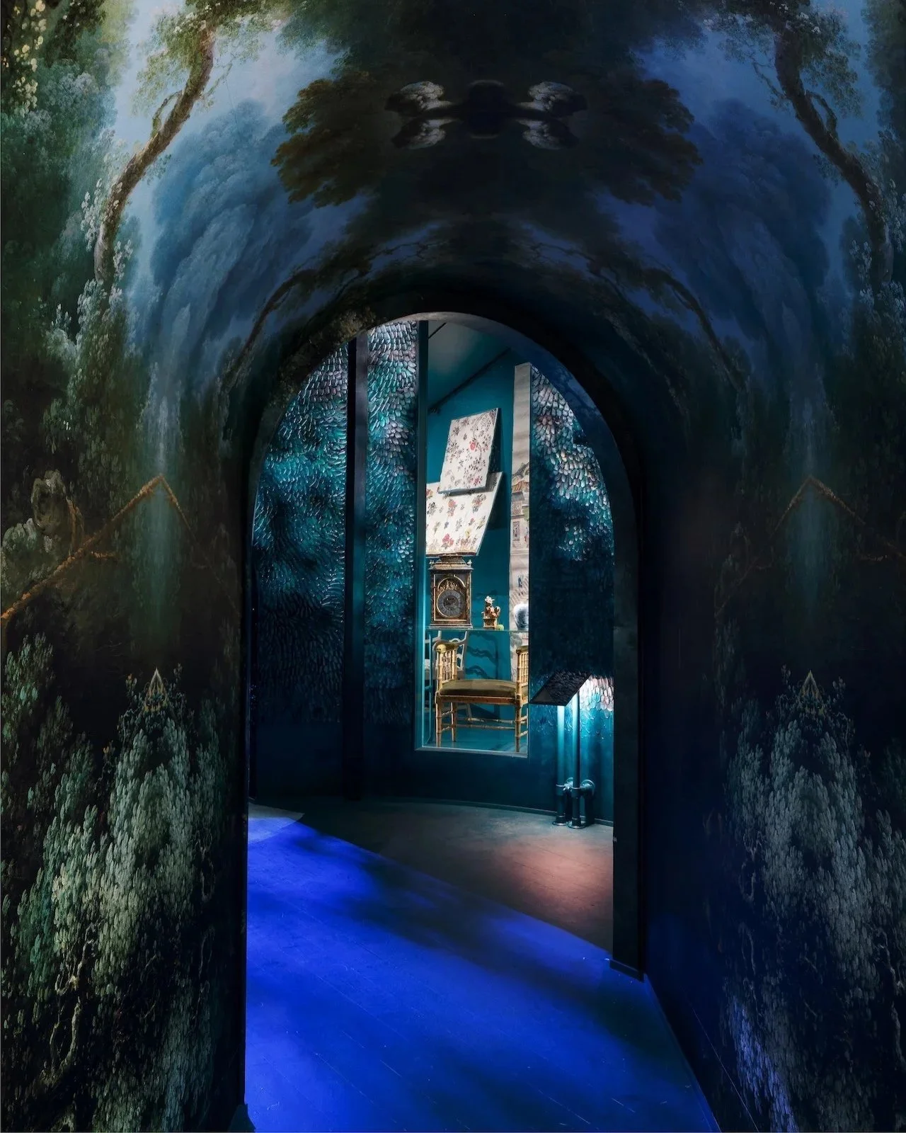 View through a decorated doorway into a room with blue walls, containing a vintage clock, floral paintings, and a wooden chair.