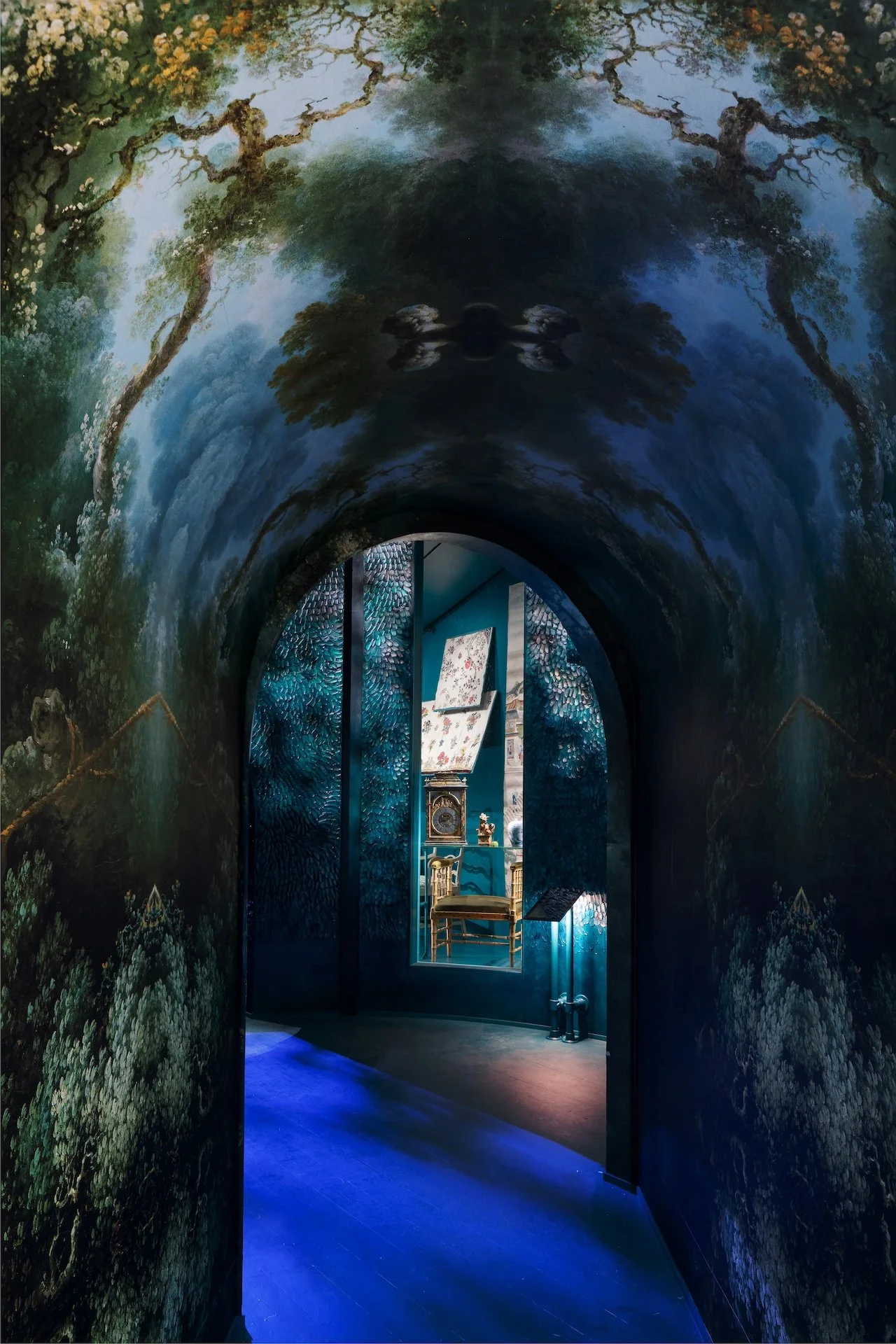 A painted and hand-sculpted plaster tunnel with a blue and green forest scene on the walls and ceiling, leading to a room with a chair, a clock, and artwork on the wall.