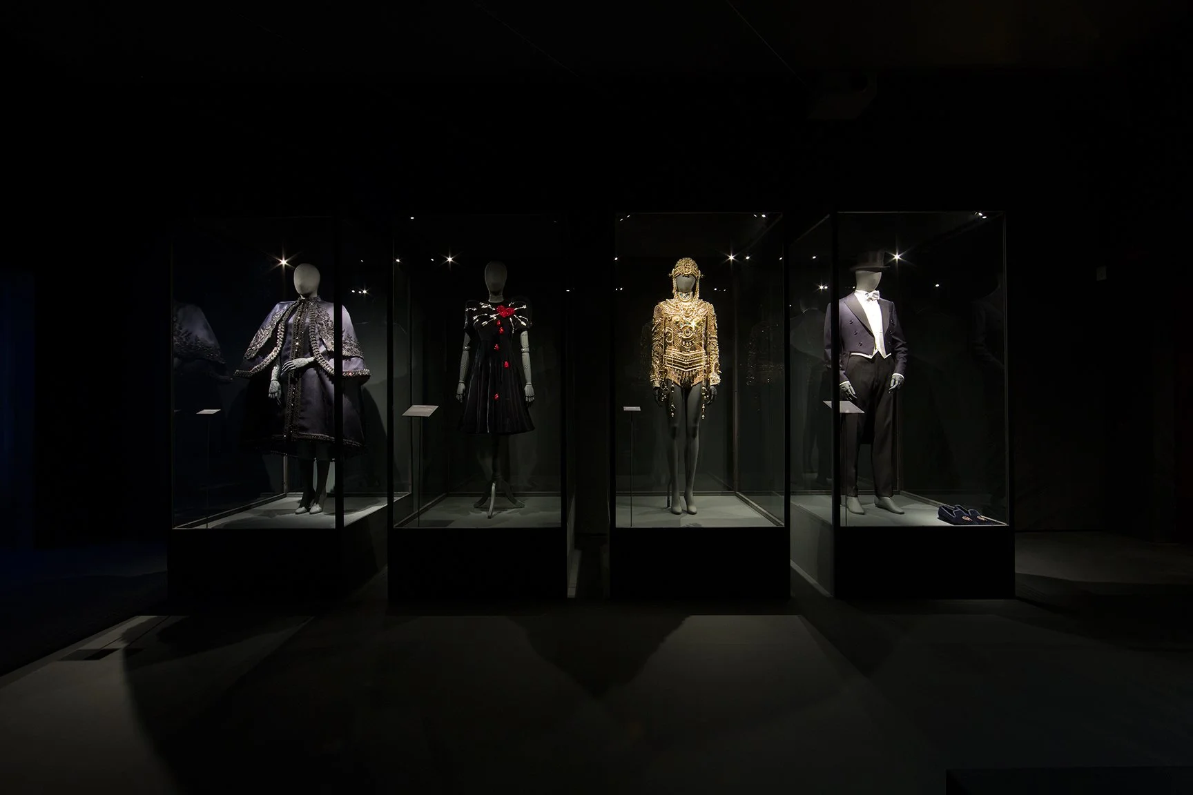 Display of five mannequins dressed in luxurious, ornate costumes in a dark exhibition space with spotlights highlighting each outfit. With hand-carved mannequins.