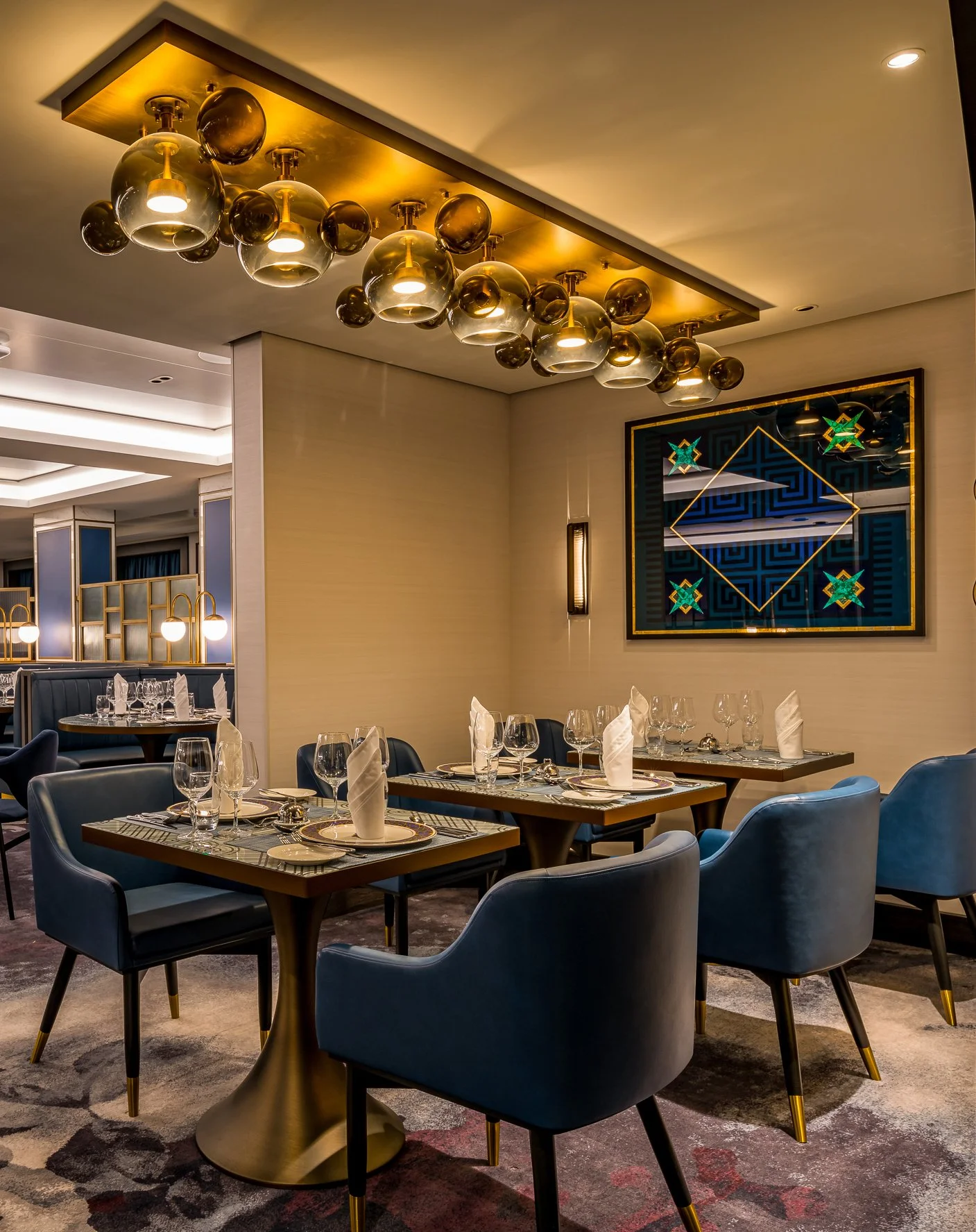 Elegant restaurant dining area with tables set with white napkins and glassware, modern blue chairs, a decorative gold and black chandelier, and a colorful gilded glass geometric wall art.