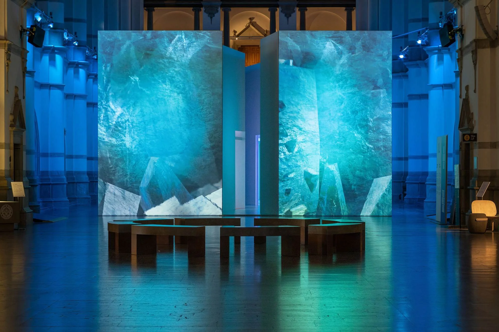 An indoor art installation featuring large, standing panels displaying a dynamic, icy blue aquatic scene, illuminated with blue light, within a grand museum with classical architectural elements and wooden flooring.