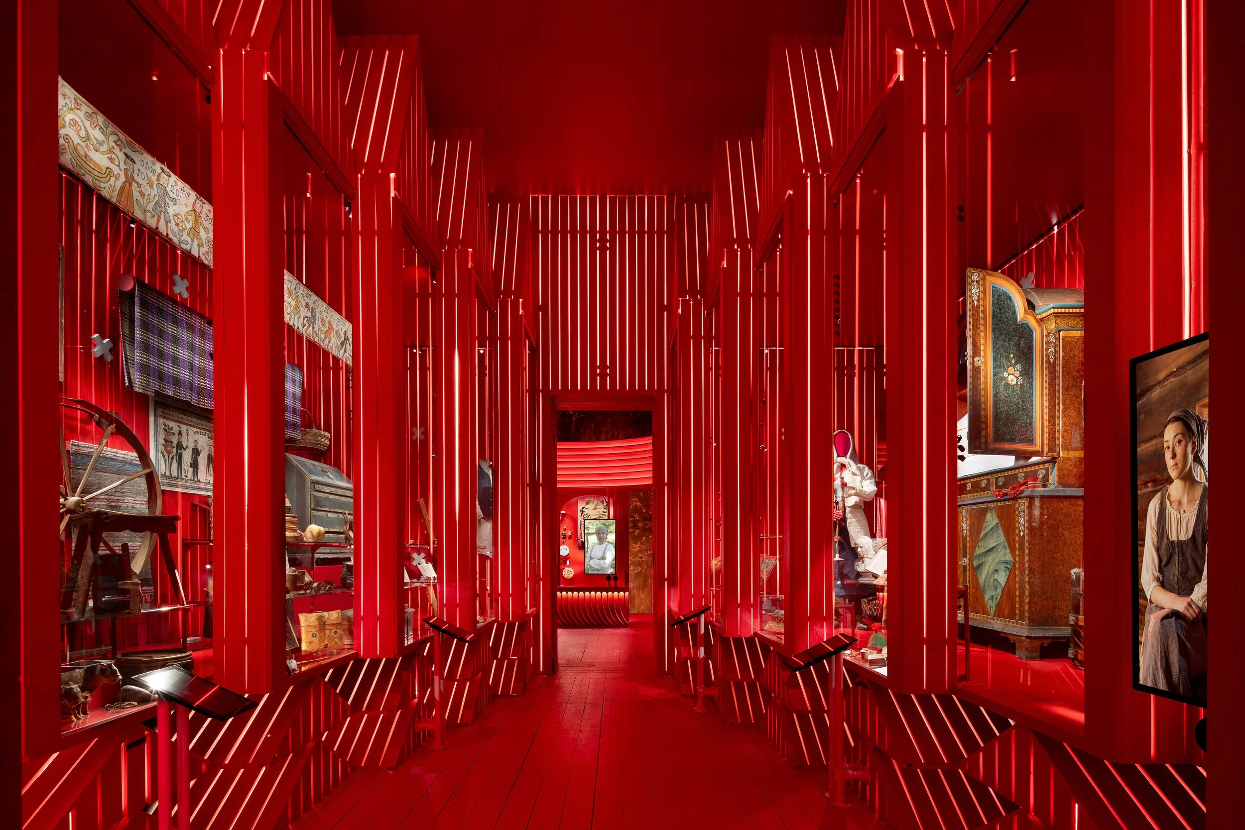 A vibrant interior space featuring red vertical slats with integrated lighting, various displayed art pieces, and furniture, creating a modern, artistic atmosphere.