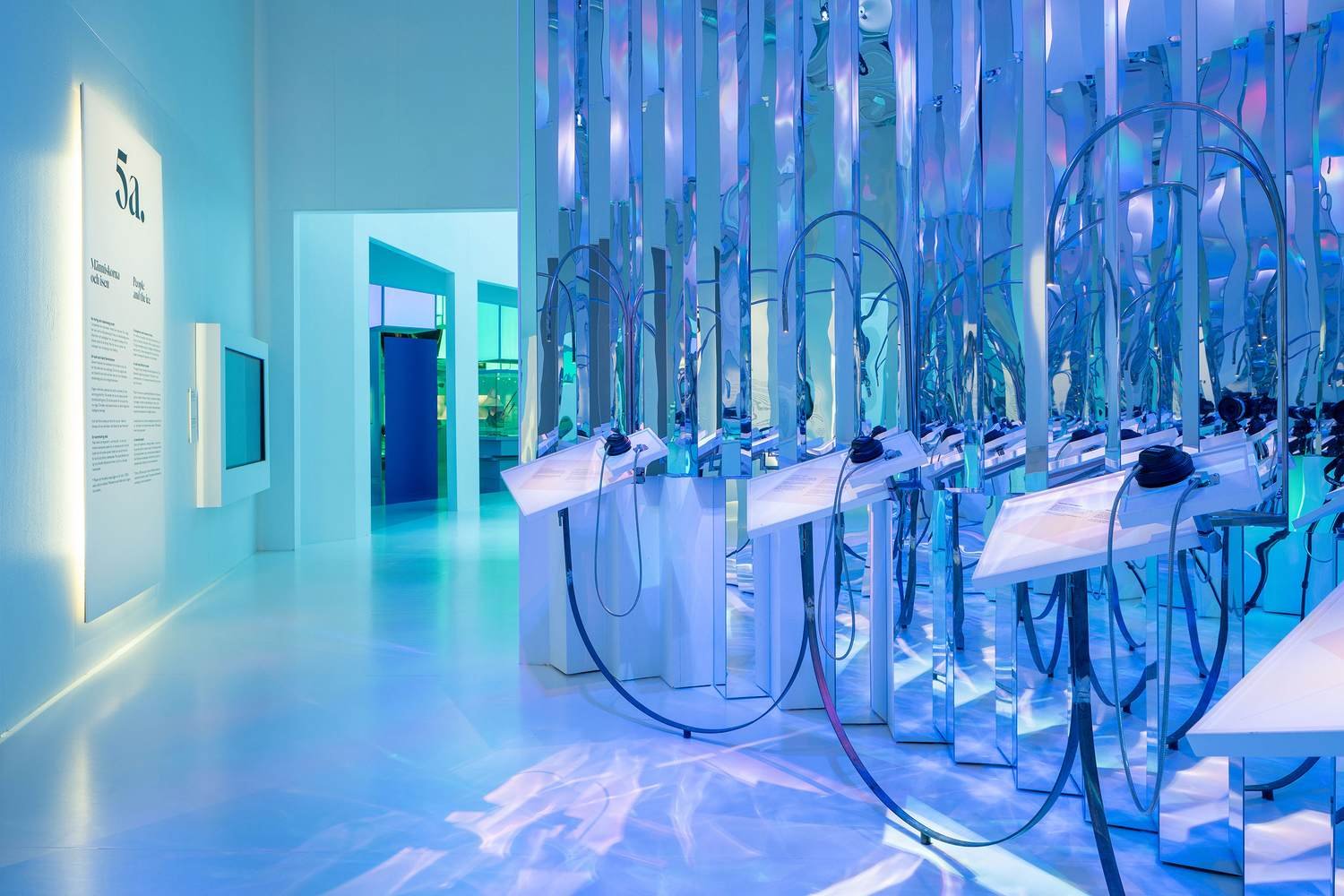 A modern art installation with blue and purple lighting, featuring a row of transparent display stands with headphones, set against a background of reflective, curved metal structures and walls with informational panels.